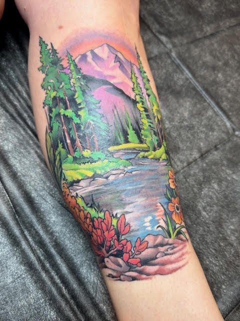 Colorful realistic tattoo of a mountain landscape with trees, a river, flowers, and a sunrise or sunset on a forearm.