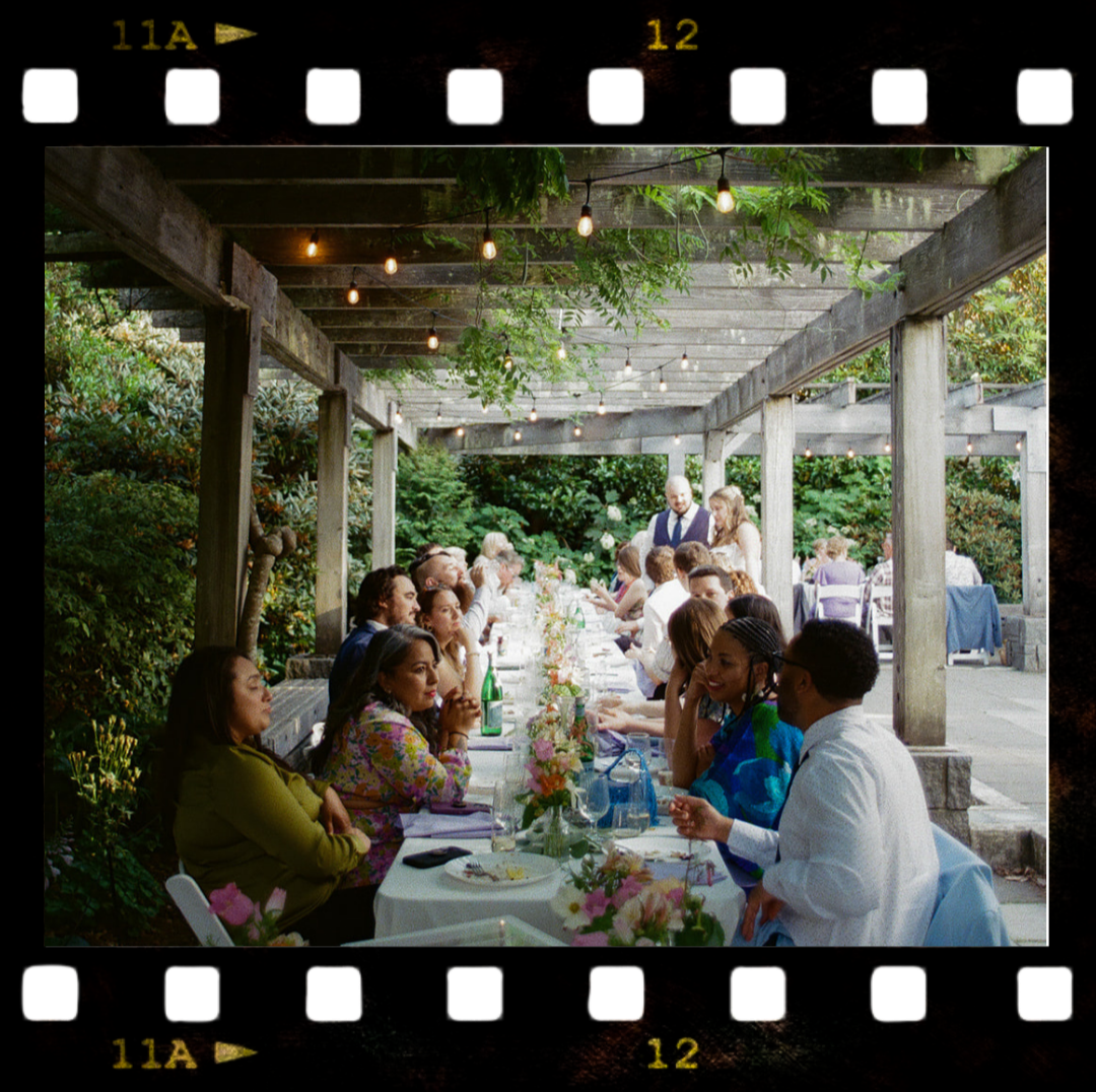 People sitting at a long outdoor table decorated with flowers and bottles, enjoying a meal under string lights at a garden wedding reception.