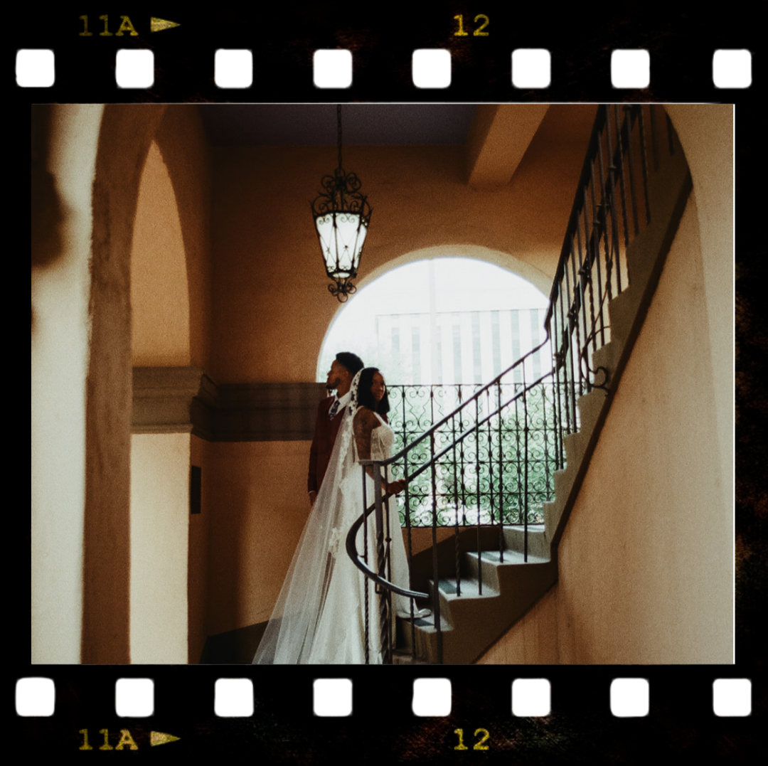 A bride and groom standing on a staircase inside a building with arched windows and a hanging lantern, captured through an overlaid film strip border.
