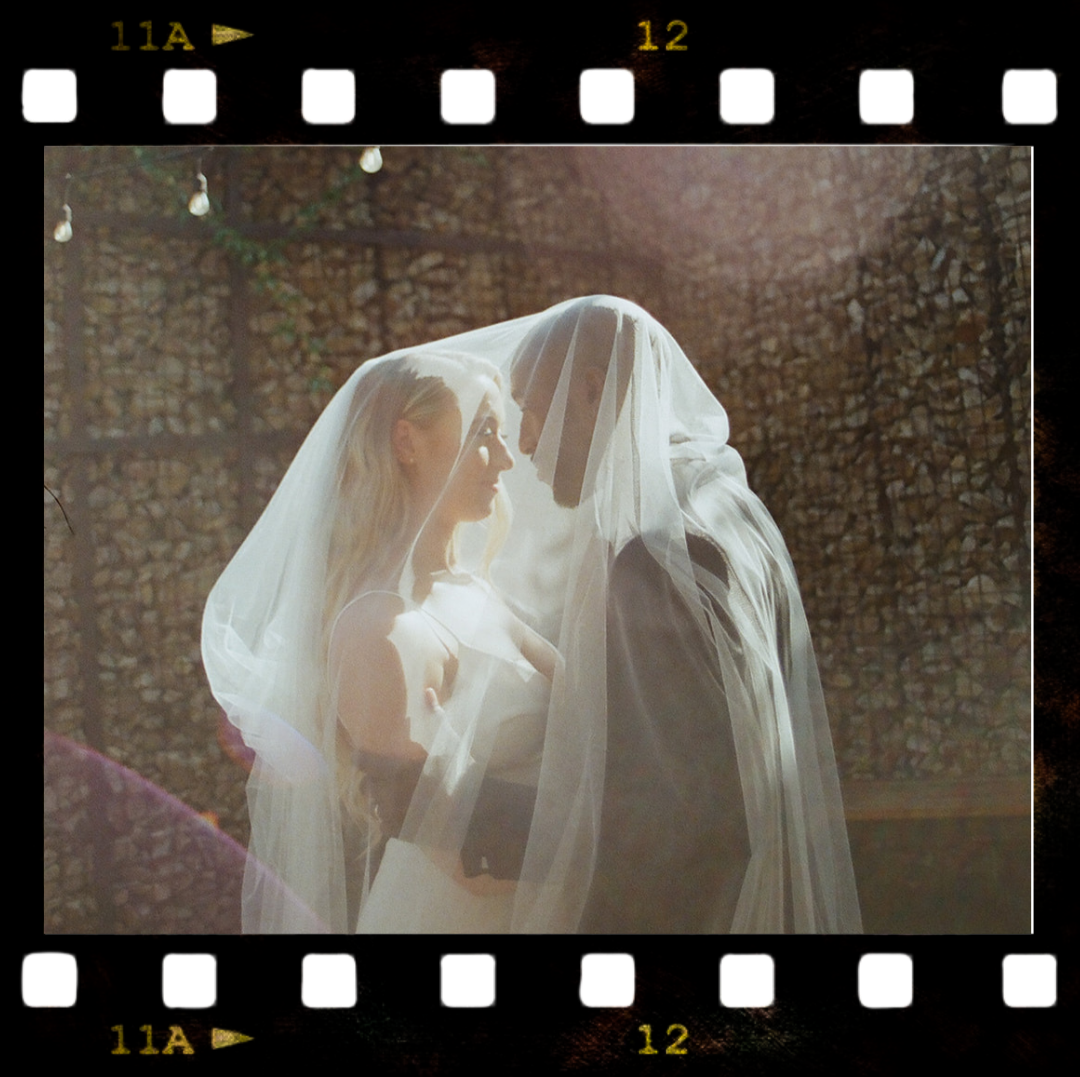 A bride and groom face to face under a sheer veil, standing outdoors in front of a woodstack, with sunlight creating a warm glow.