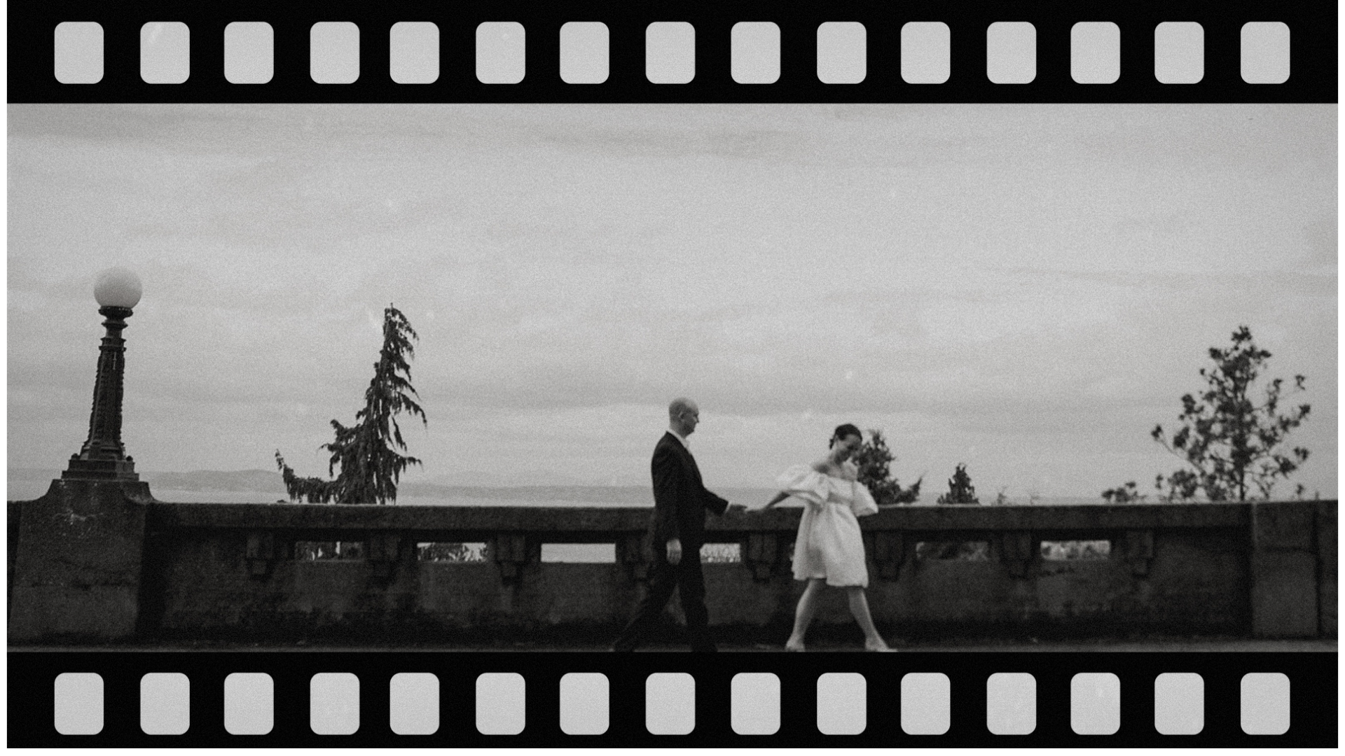 Black and white photo of a couple in vintage clothing walking along a bridge or balcony with decorative railing indoors or on a terrace, with trees and a lamp post in the background.