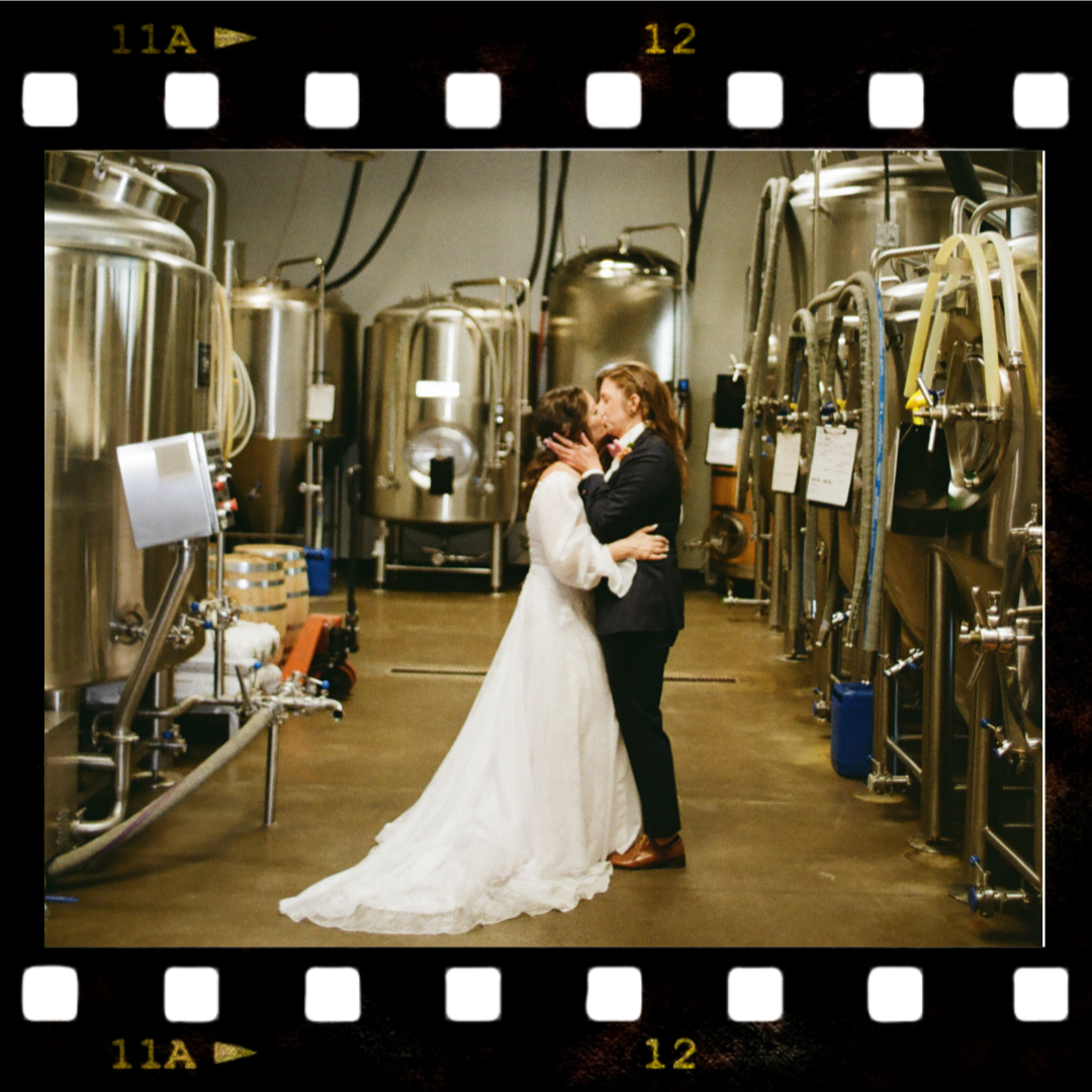 Two women in wedding attire sharing a kiss in a brewery or industrial beverage facility with stainless steel tanks and equipment in the background.