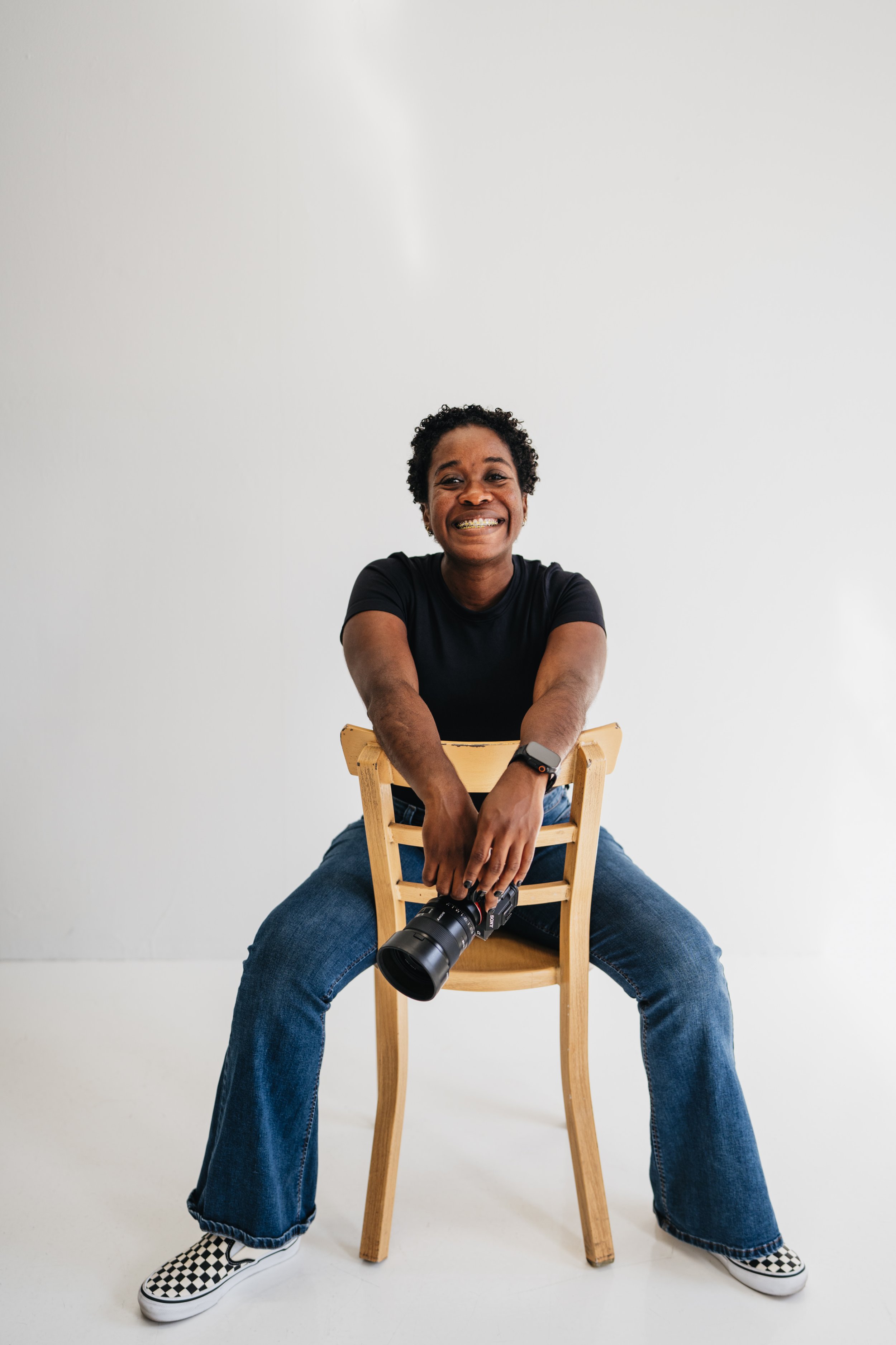 Person sitting on a wooden chair, smiling, with a camera hanging between their legs, dressed in a black t-shirt, blue jeans, and checkerboard Vans sneakers, against a plain white background.