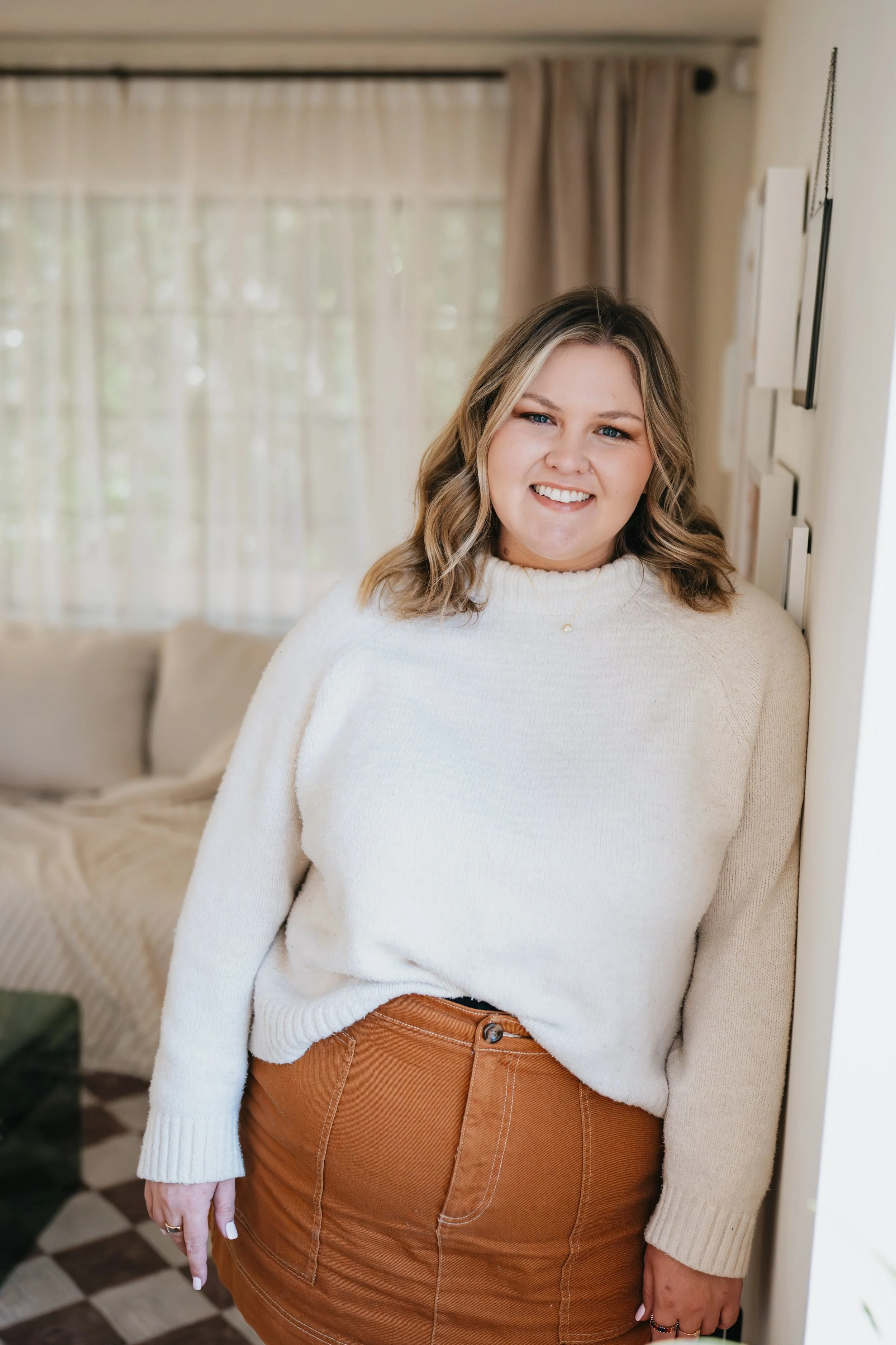 A woman with wavy blonde hair smiling, wearing a cream-colored sweater and a brown skirt, standing indoors by a white wall with framed pictures. There is a window with beige curtains in the background.