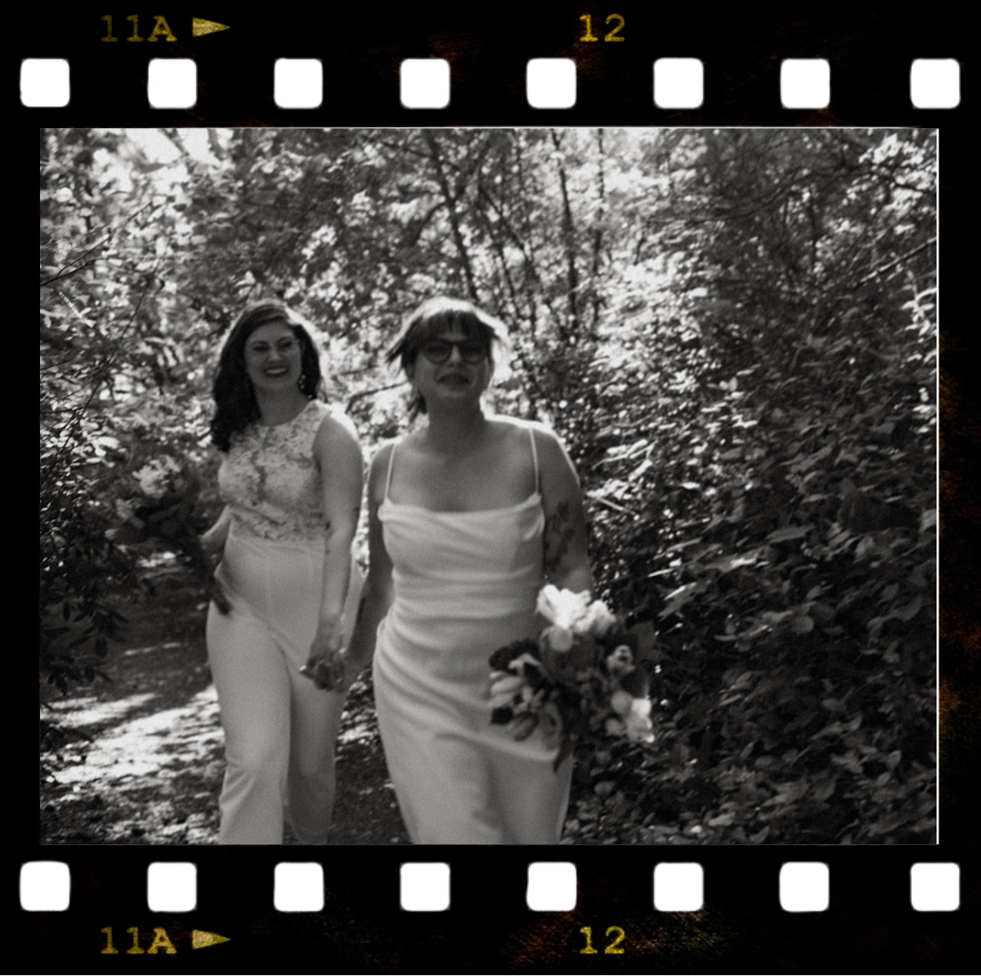 Two women in wedding dresses walking hand in hand outdoors through a wooded area, one smiling and holding a bouquet, in black and white.