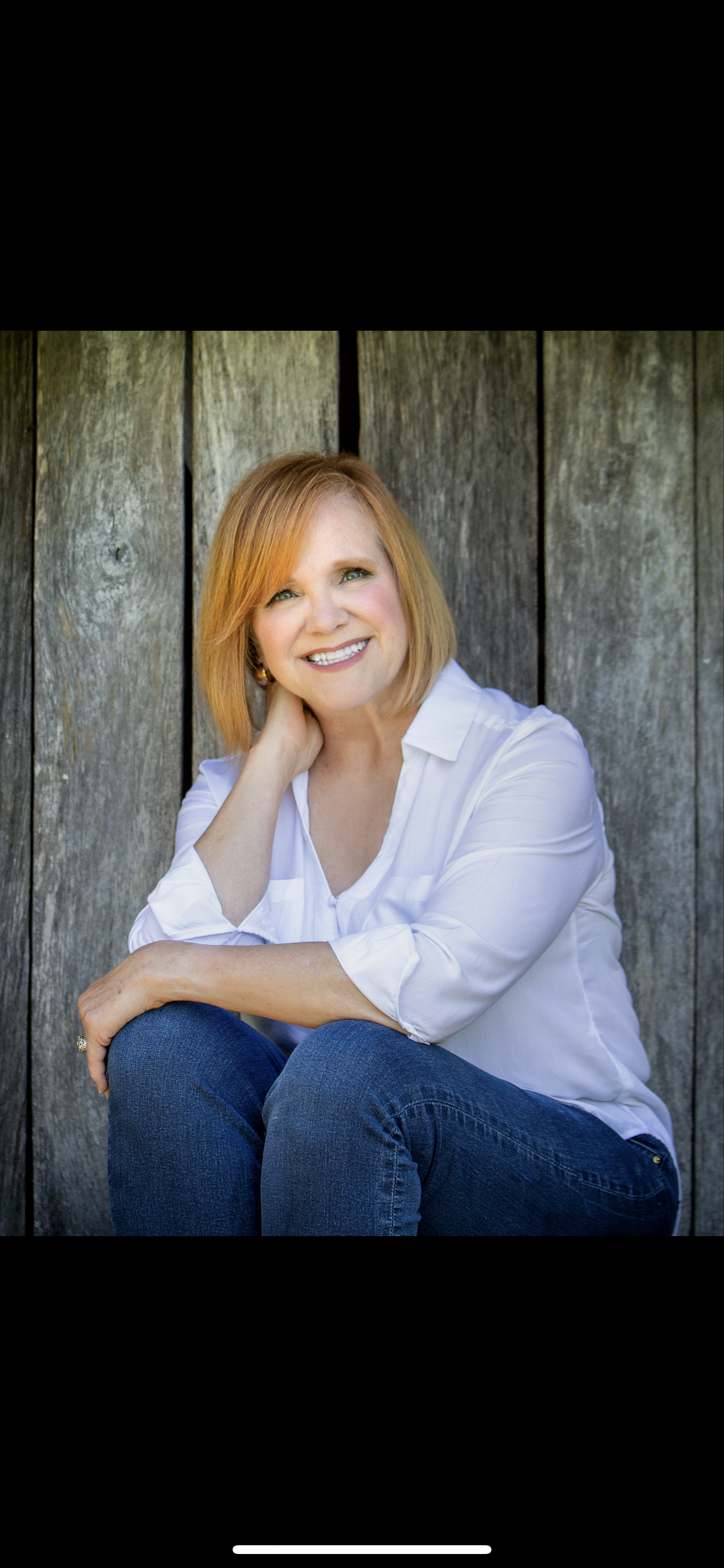 Person with short red hair smiling, wearing a white shirt and jeans, sitting in front of a wooden background.