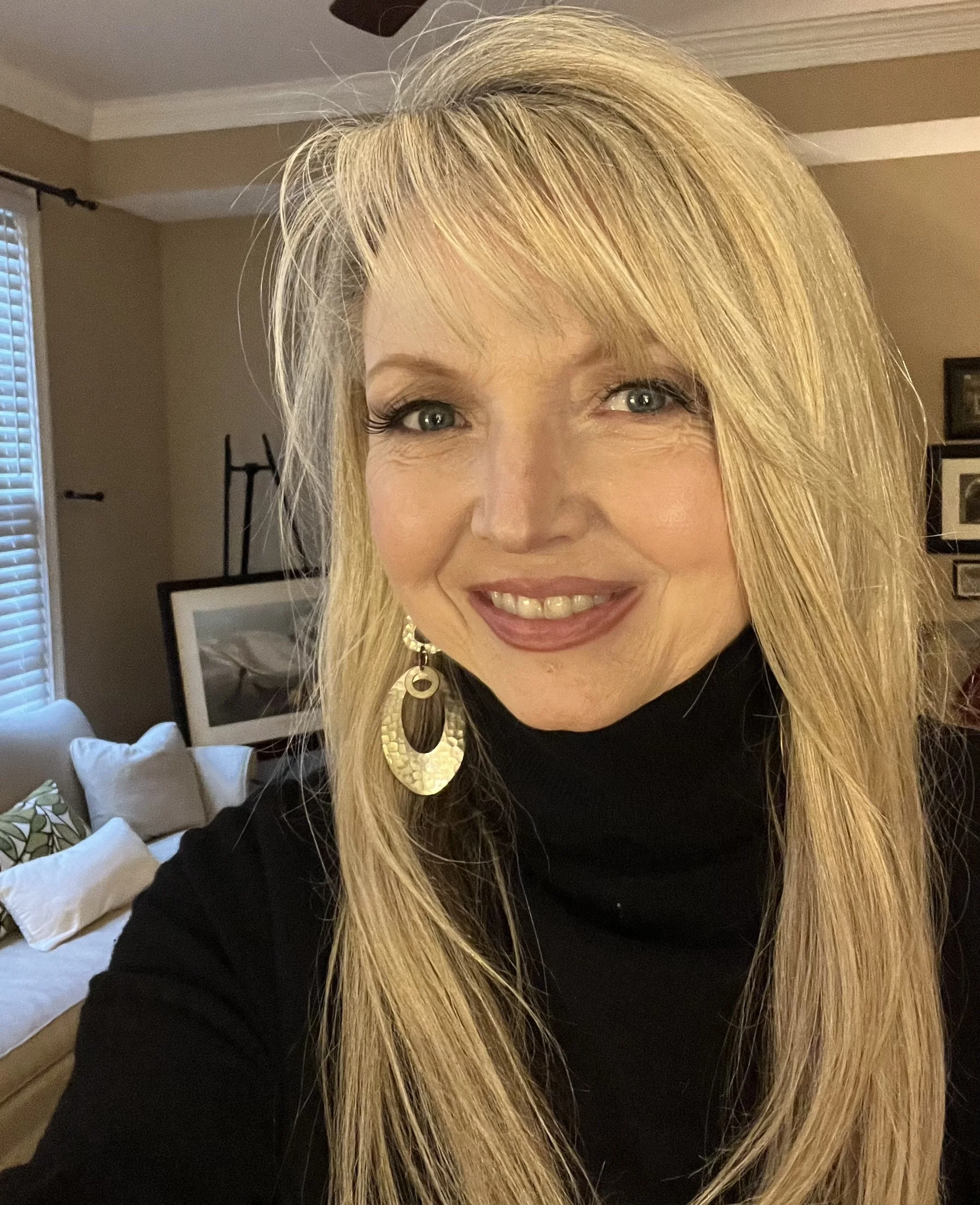 A woman with long blonde hair, wearing a black turtleneck and large gold earrings, smiling in a living room with beige walls and framed artwork.