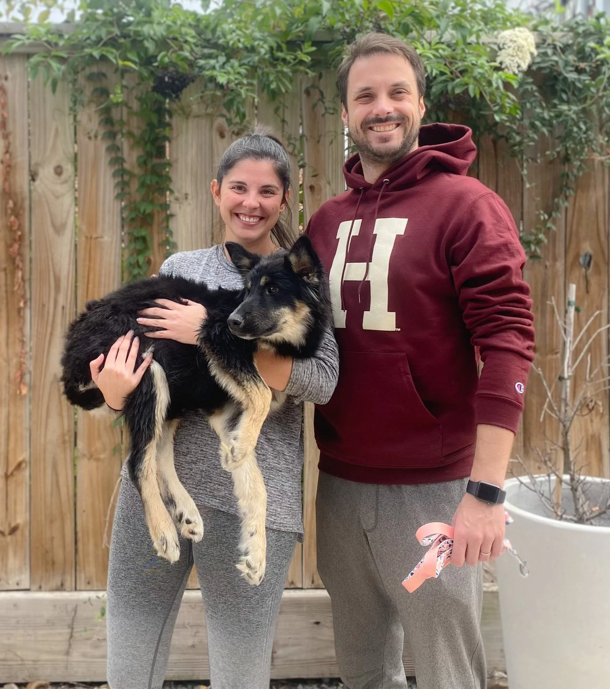 A woman and man standing outdoors in front of a wooden fence, smiling at the camera. The woman is holding a black and tan puppy. The man is wearing a maroon hoodie and has a smartwatch, holding a ribbon or leash in his hand.