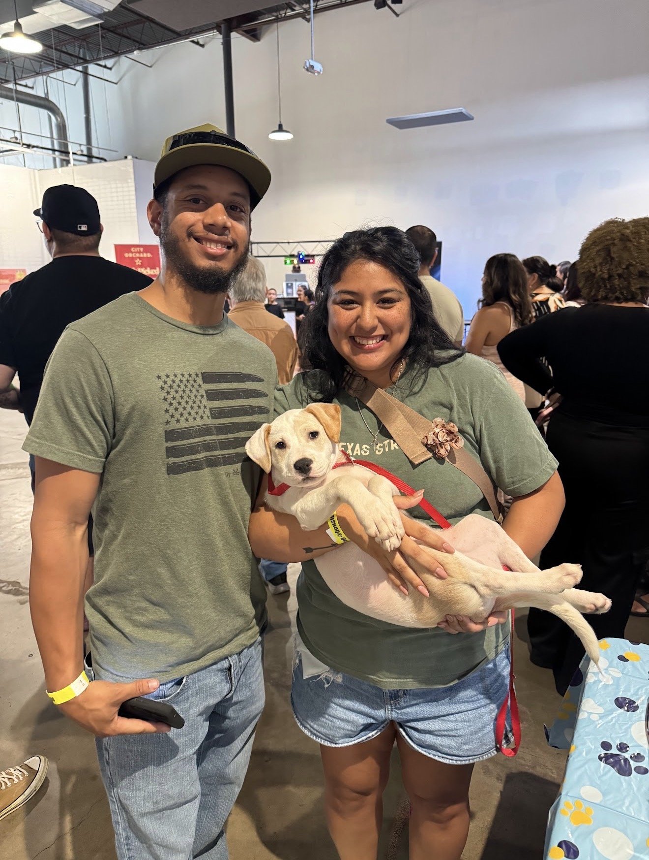 A smiling man and woman holding a puppy at an indoor event. The man is wearing a green t-shirt and a cap, and the woman is wearing a green t-shirt and denim shorts. The puppy is light-colored with a red collar and has a yellow wristband. There are many people in the background inside a large, well-lit industrial-style space.