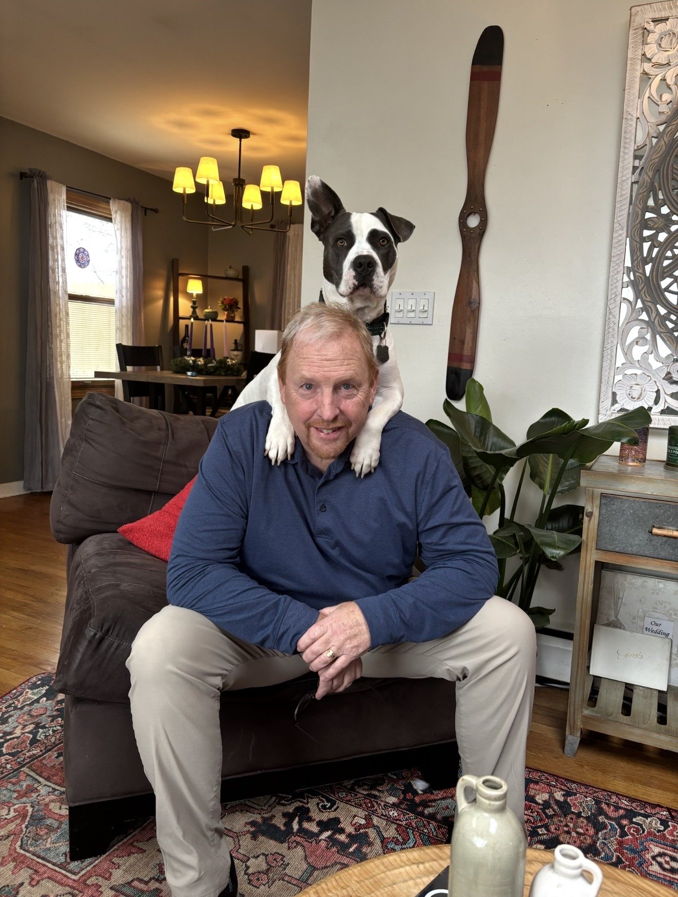 A man sitting on a couch with a dog standing behind him, with the dog's paws on the man's shoulders in a living room.