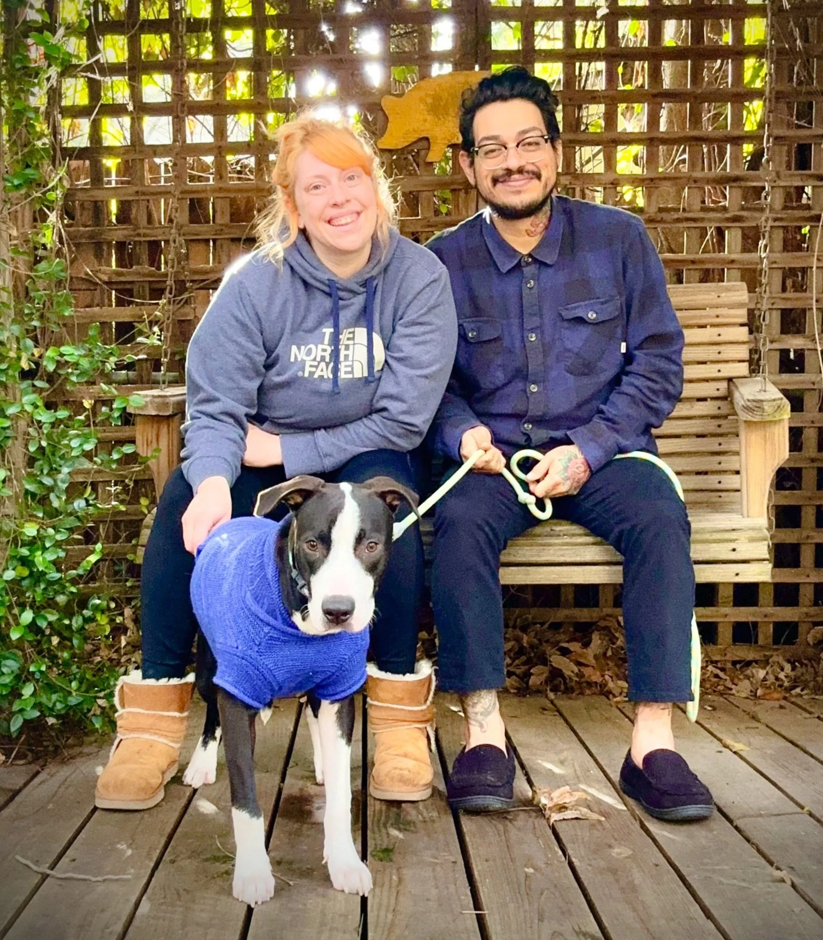 Two smiling people, a woman with red hair wearing a gray North Face hoodie and a man with glasses, a beard, and tattoos wearing a navy blue shirt, are sitting on a wooden bench outdoors. They are accompanied by a black and white dog wearing a blue sweater. The background features wooden fencing, greenery, and sunlight filtering through.