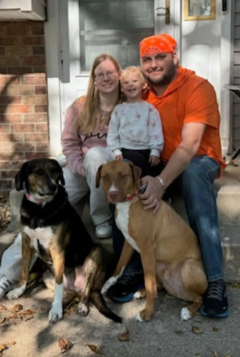 A family of three with two dogs sitting outside in front of a house, smiling at the camera.