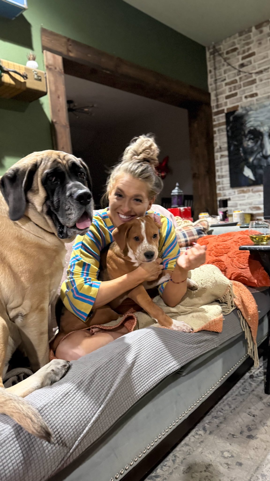 A woman sitting on a couch with two dogs, one large and one small, smiling at the camera. The large dog is sitting to her left, and the small dog is on her lap. The room has exposed brick and wooden decor, with a painting of a dog on the wall.