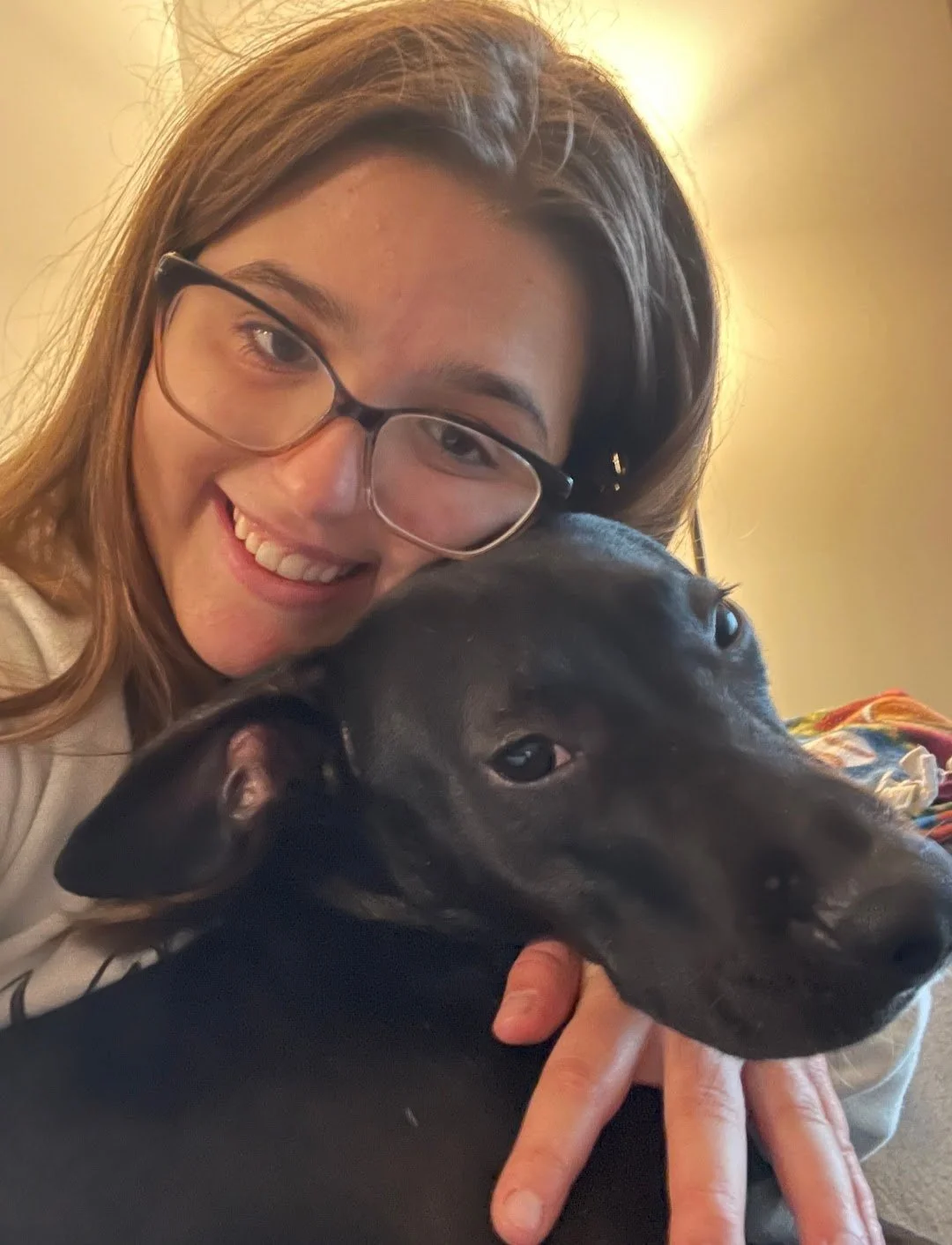 A woman with glasses smiling and taking a selfie with a black dog lying close to her face.