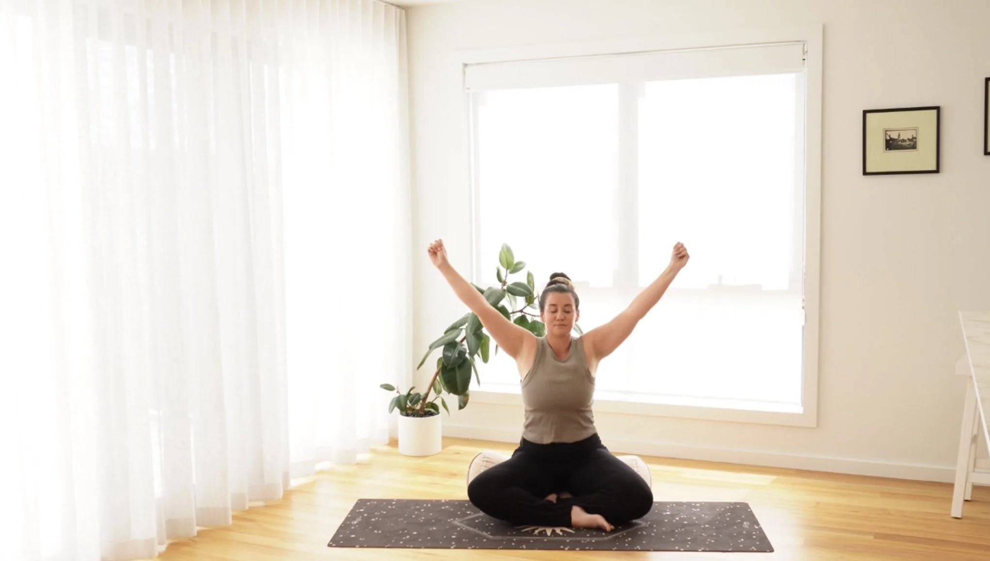 Woman practicing yoga in a bright, airy room with large window, sheer curtains, potted plant, and framed pictures on wall.