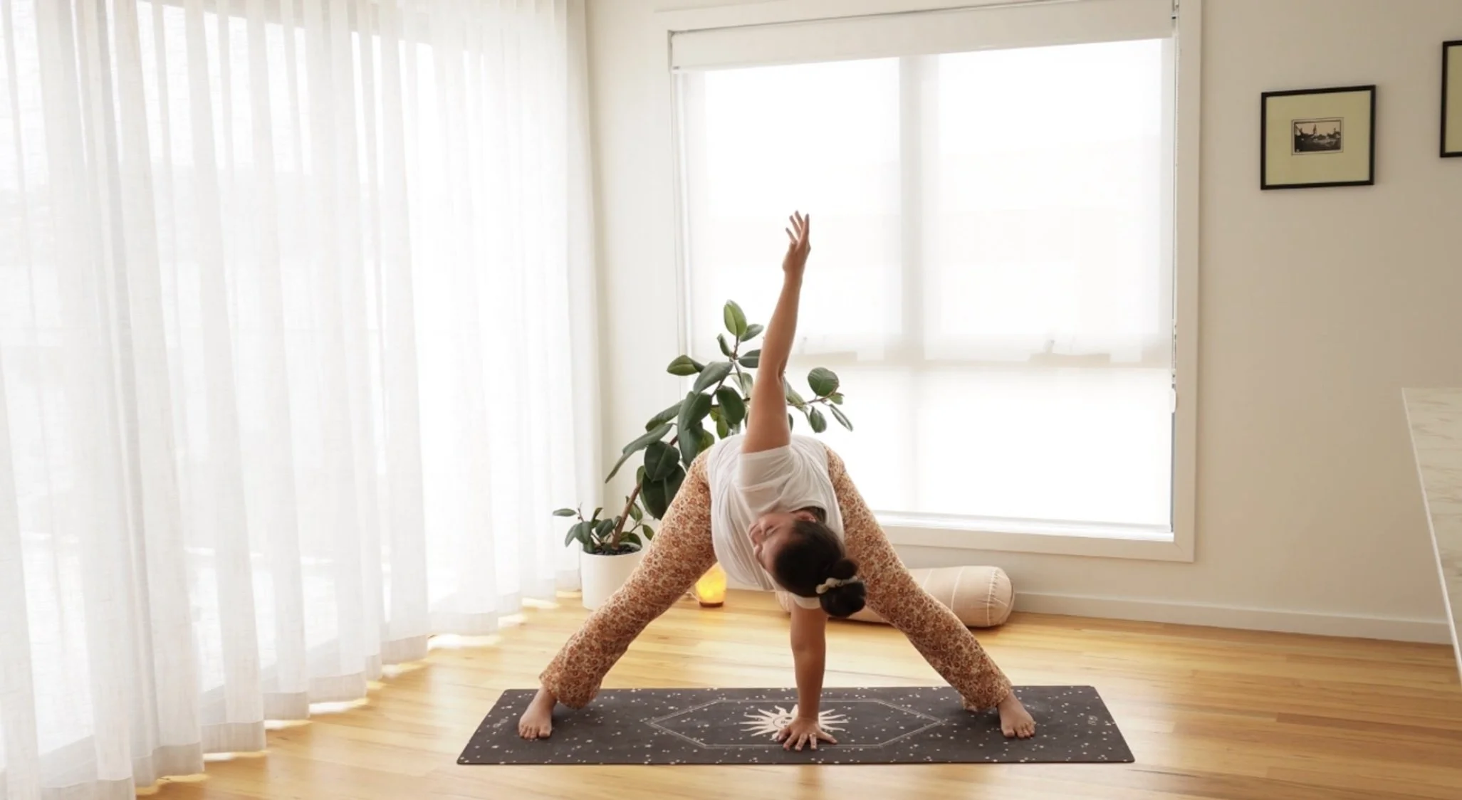 A woman practicing yoga in a bright room, performing a triangle pose on a black yoga mat with a sun design, next to a tall plant, with sheer white curtains and sunlight filtering through the window.