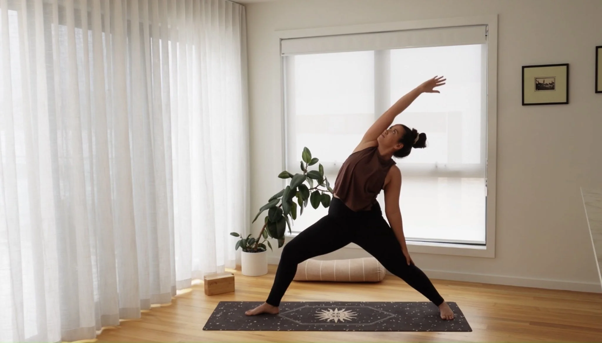 Woman practicing yoga indoors, standing in a wide-legged stance with her right arm extended overhead and her left hand on her left knee, on a black yoga mat with a sun design.