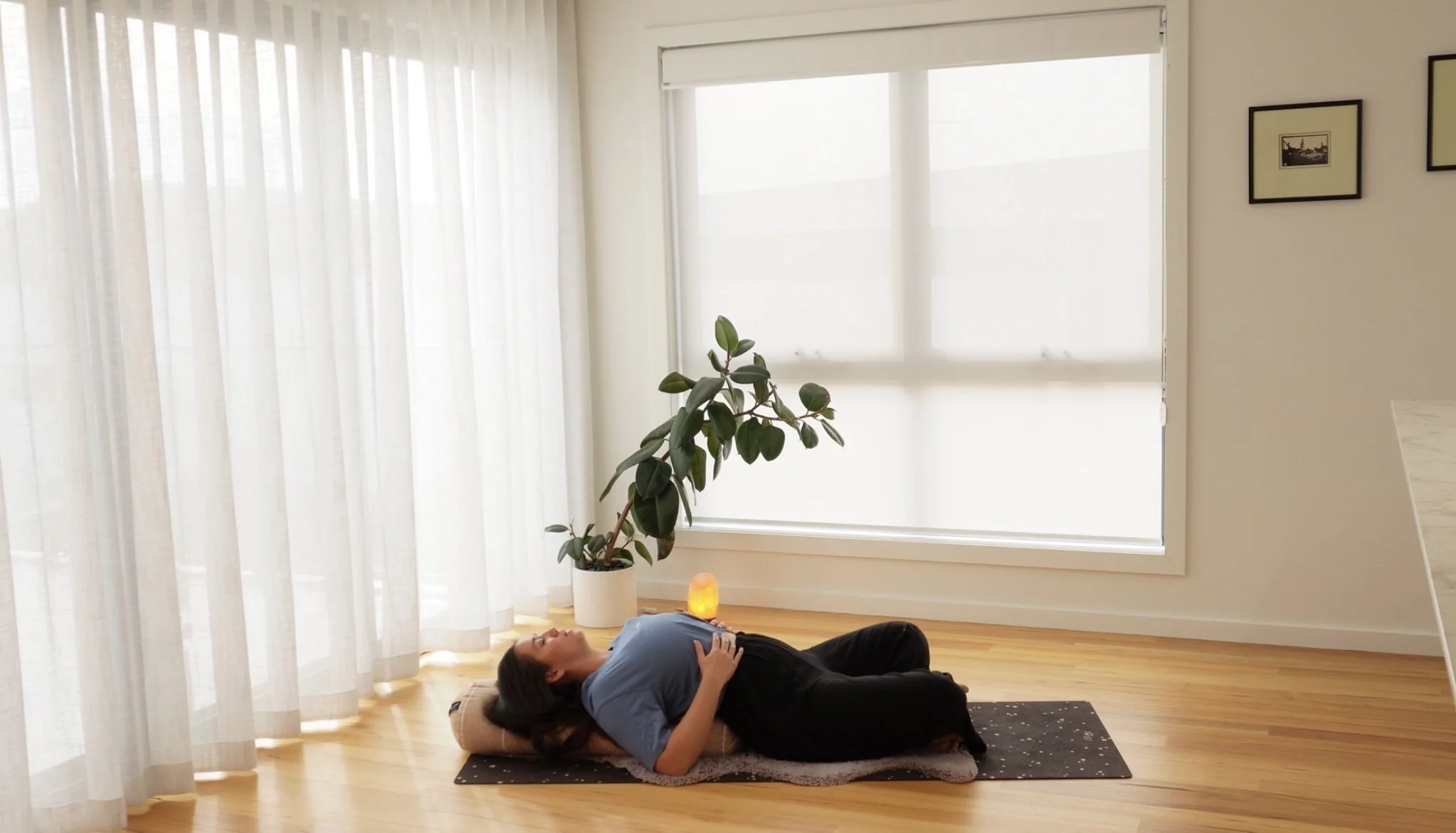 A woman lying on her back on a yoga mat, relaxing in a peaceful room with sheer white curtains, a large window, a tall potted plant, and a salt lamp.