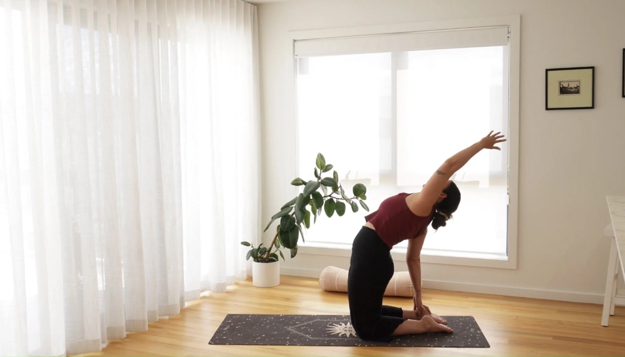 A woman practicing yoga in a bright room with wood flooring, sheer curtains, a potted plant, and framed pictures. She is on a black yoga mat, kneeling and stretching her right arm over her head.