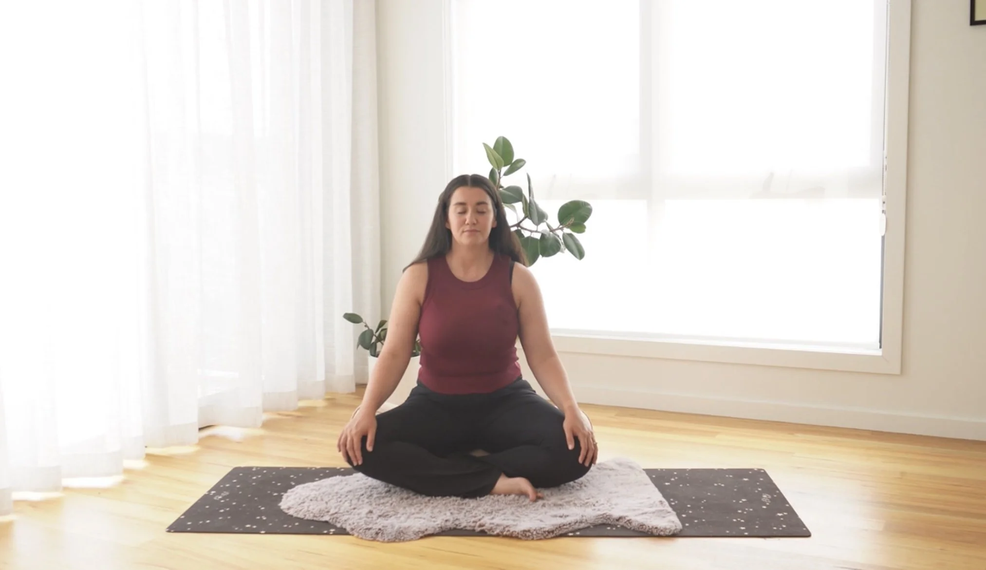 Woman meditating in a seated yoga pose on a yoga mat and plush blanket in a bright room with large windows, white curtains, and a potted plant in the background.