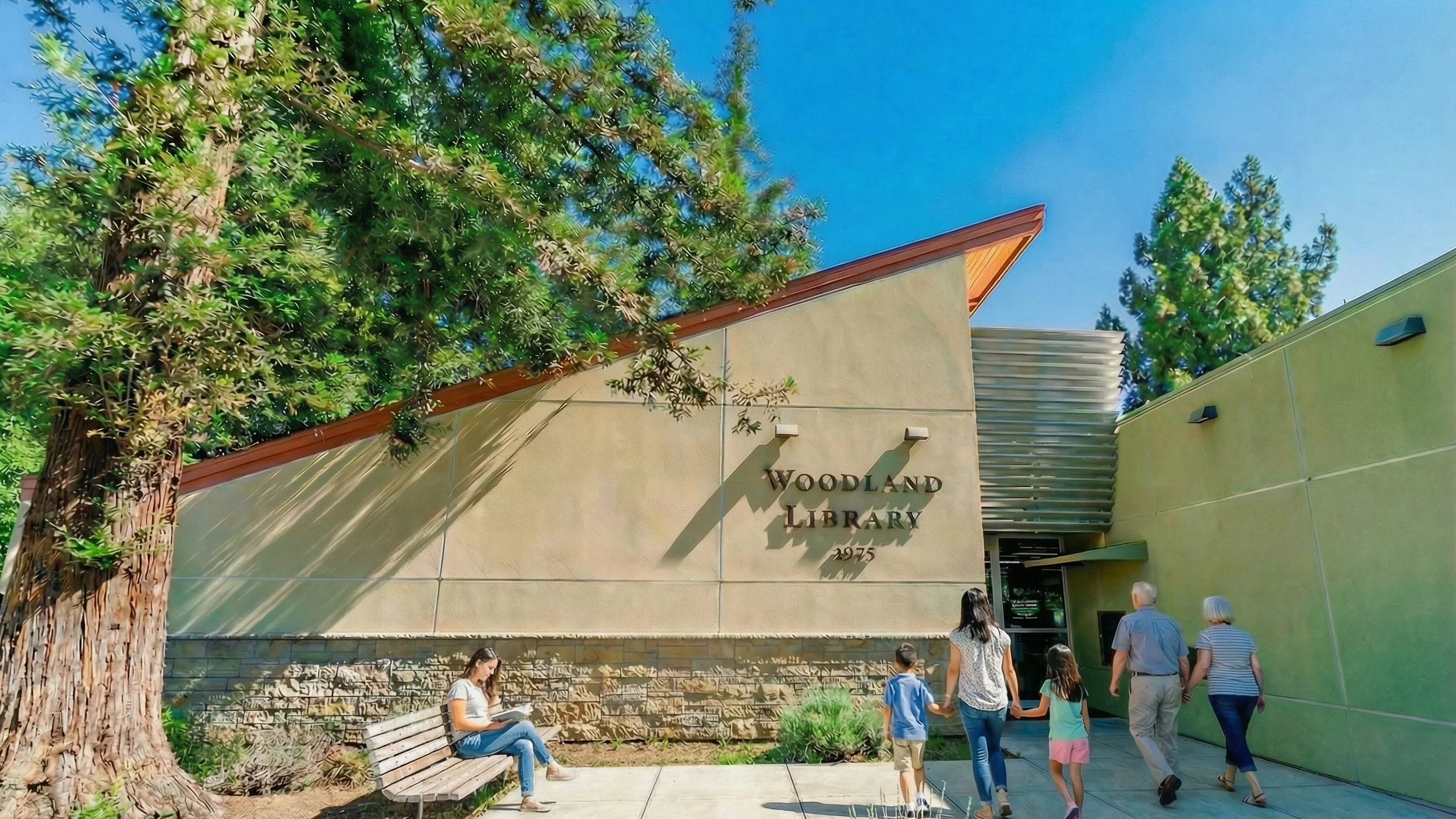 Exterior view of the Woodland Library entrance with families and children walking toward the building, surrounded by trees and landscaping.