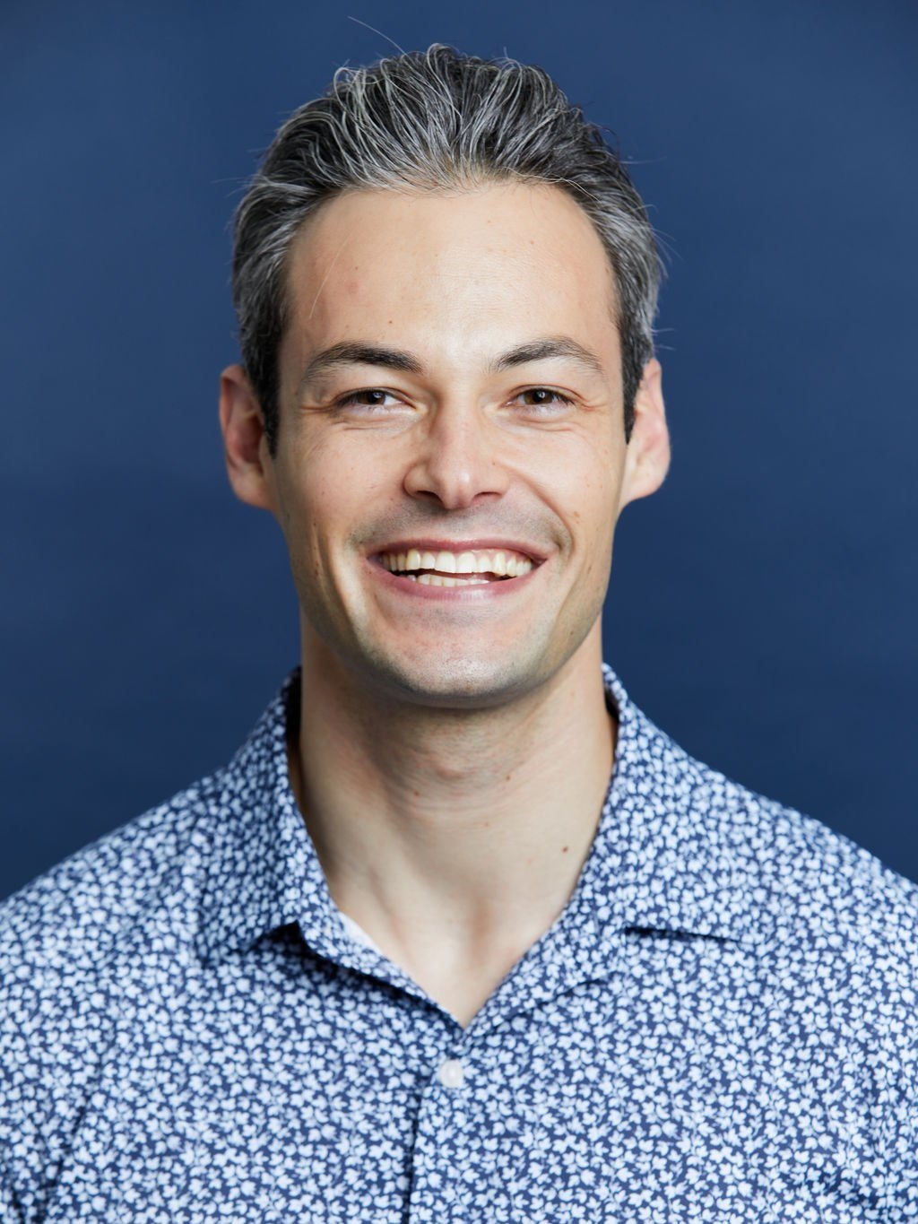 A smiling man with short dark hair, wearing a patterned shirt against a dark blue background.