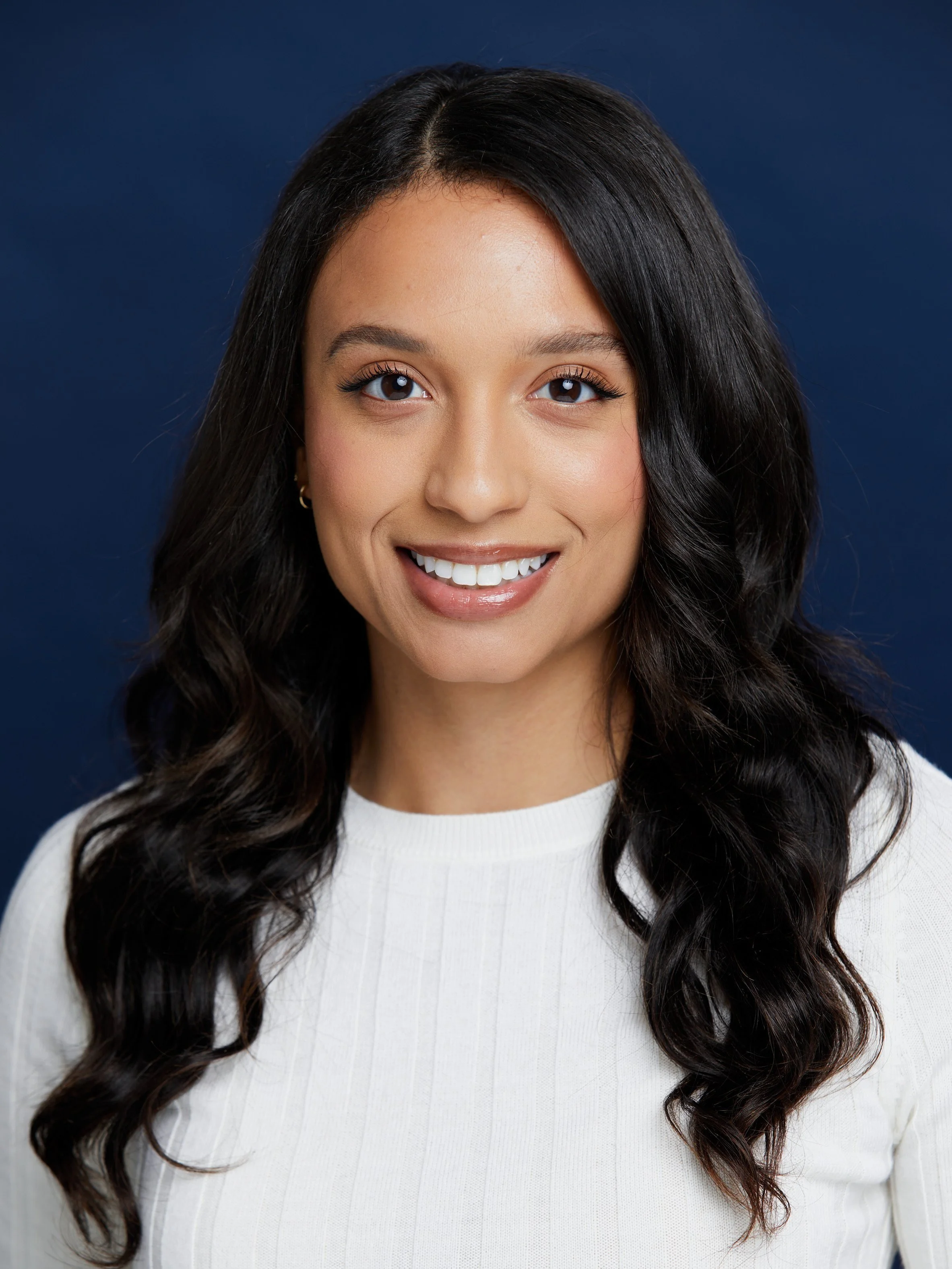 Portrait of a smiling woman with long dark wavy hair, wearing a white top, against a dark blue background.