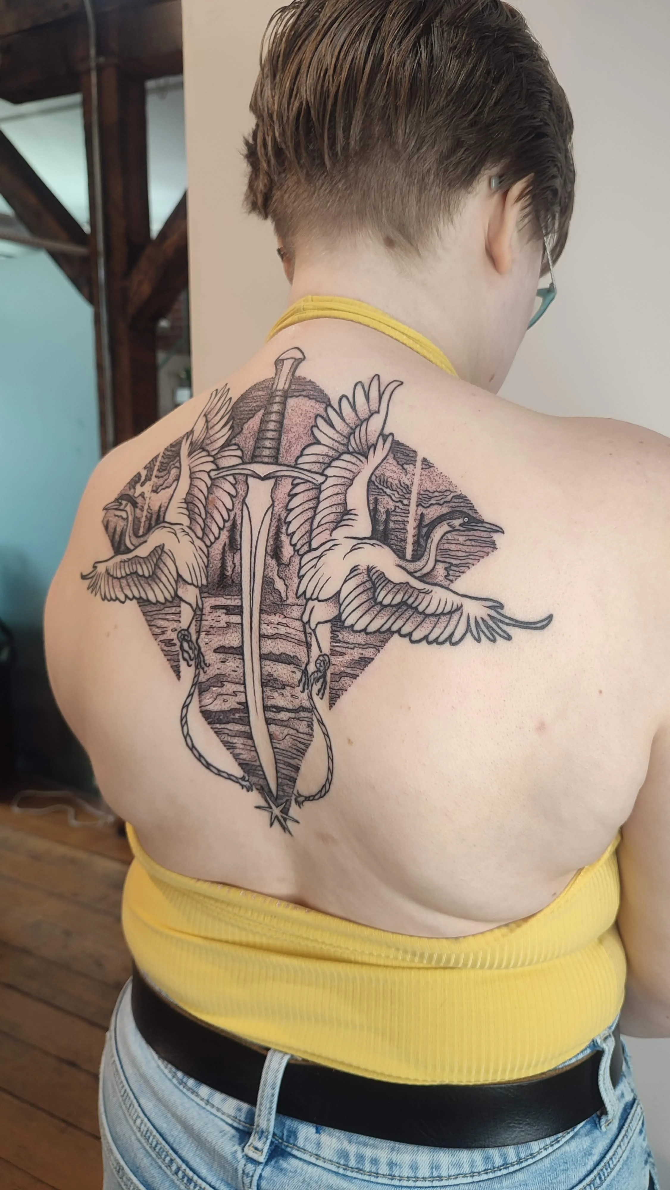 Back tattoo of two cranes with a sword and forest background on a person wearing a yellow top.