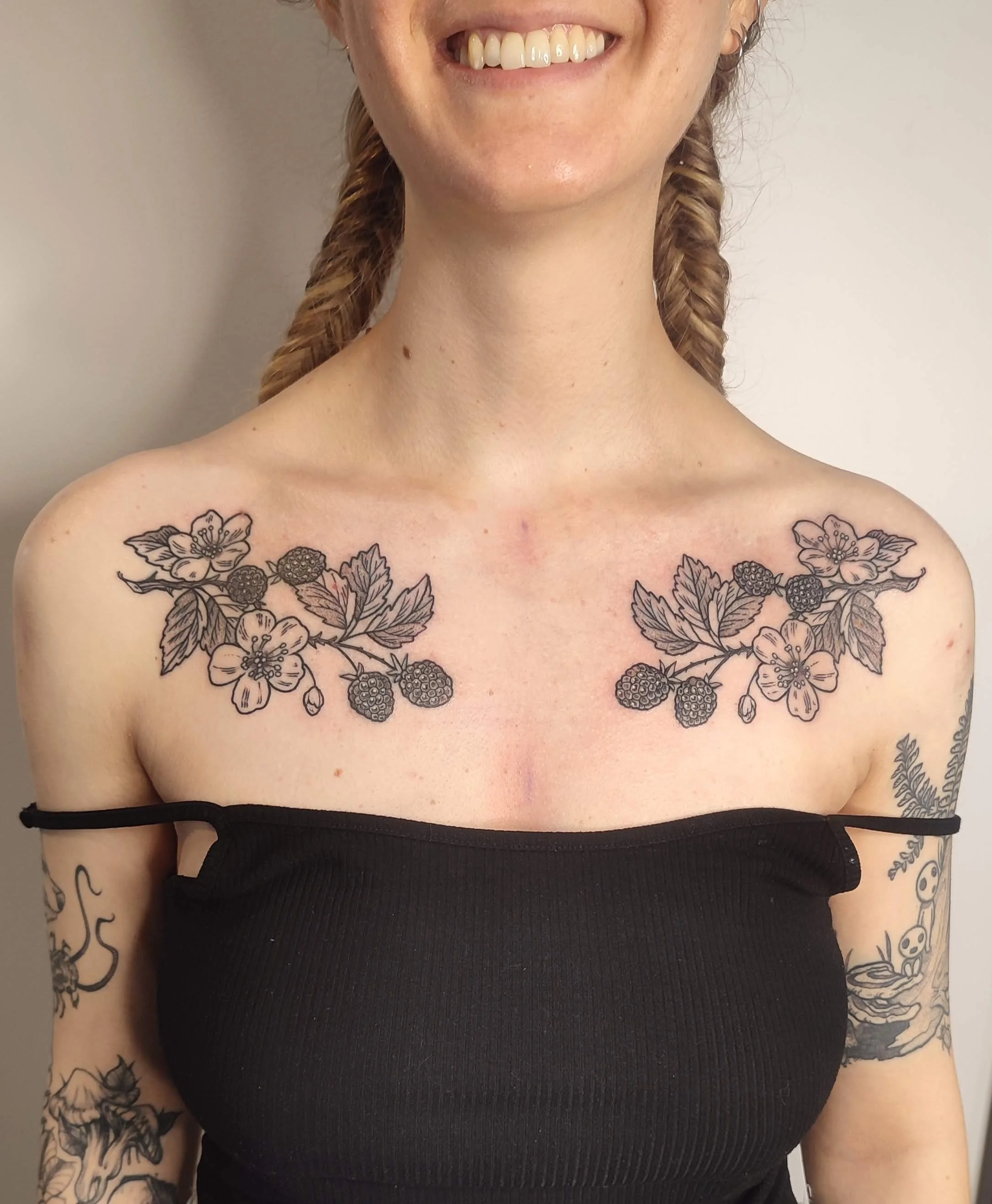 Person with floral and berry tattoos on shoulders, wearing a black top, smiling.