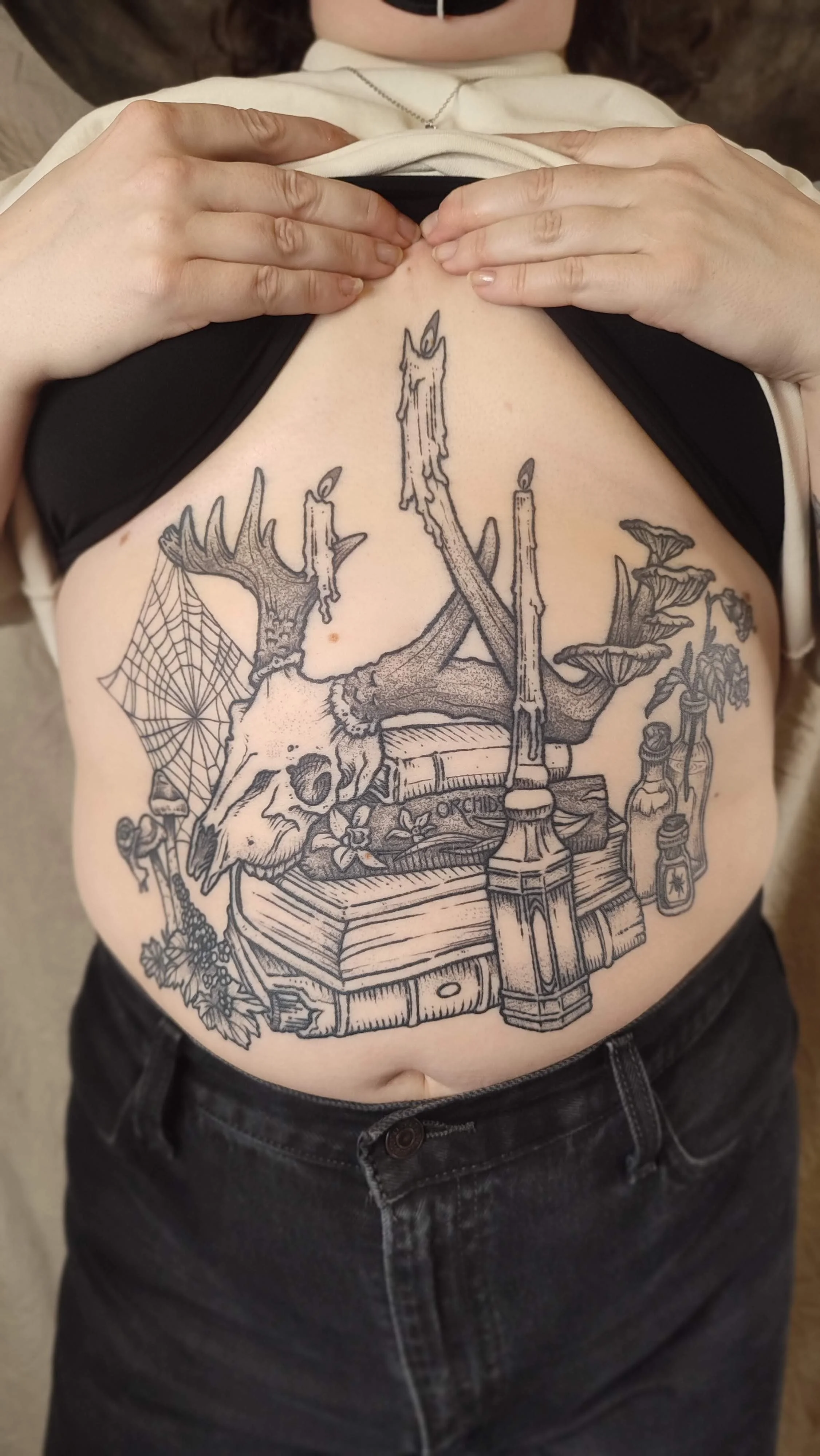 Tattoo on a person's torso featuring a deer skull with antlers, candles, books, cobweb, mushrooms, flowers, and potion bottles.