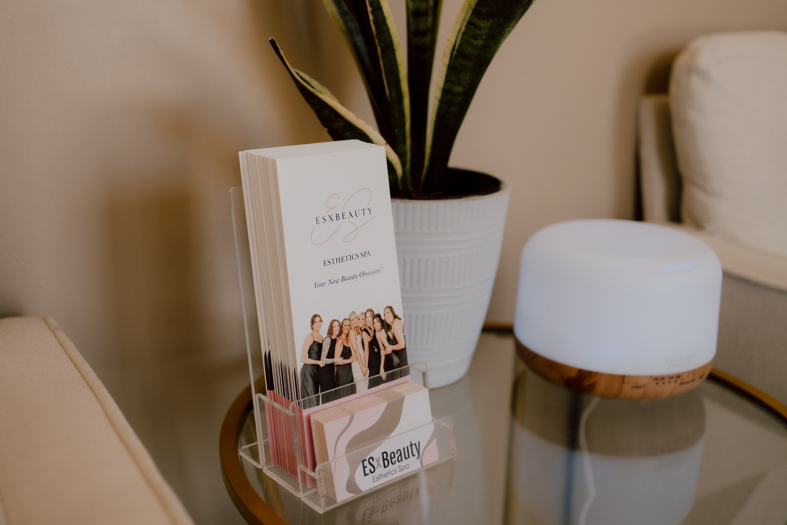 Brochures for ESX Beauty Esthetics Spa on a glass table, with a large potted plant behind, and a white smart diffuser on a wooden base to the right.