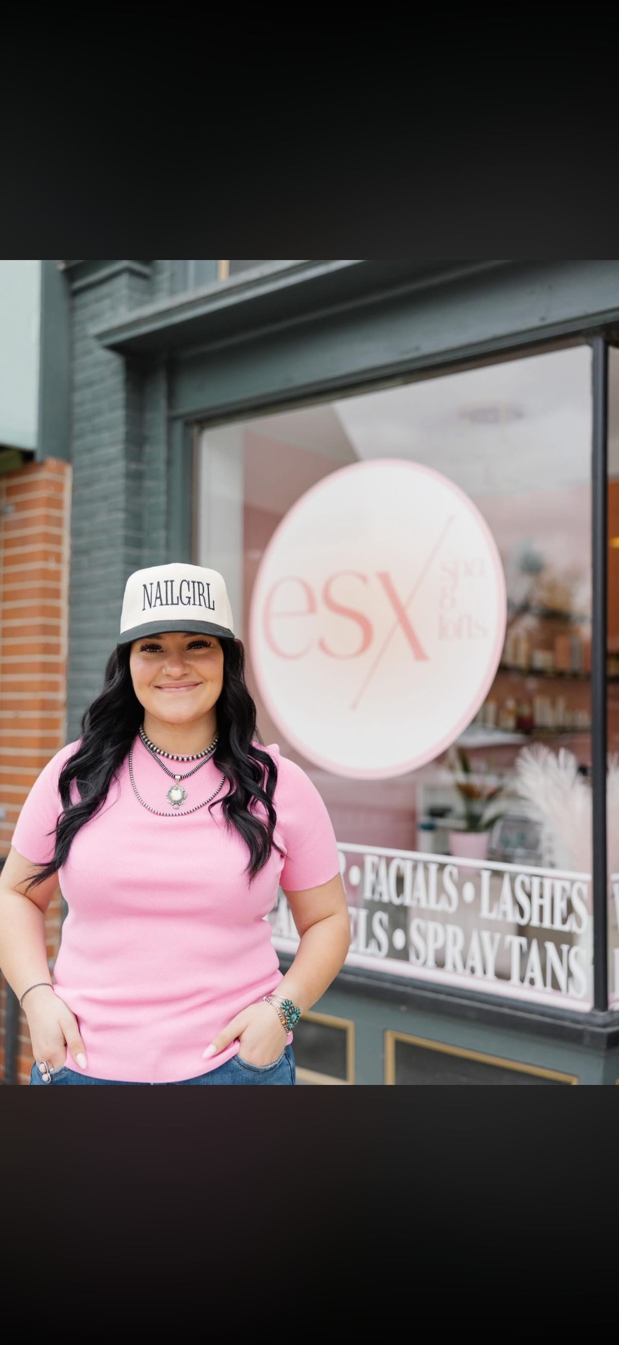 A woman with long dark hair wearing a pink shirt, a black and white hat that says 'NAILGIRL,' and jewelry stands outside a beauty salon with a large round sign reading 'esx' and services listed on the window including facials, lashes, and spray tans.