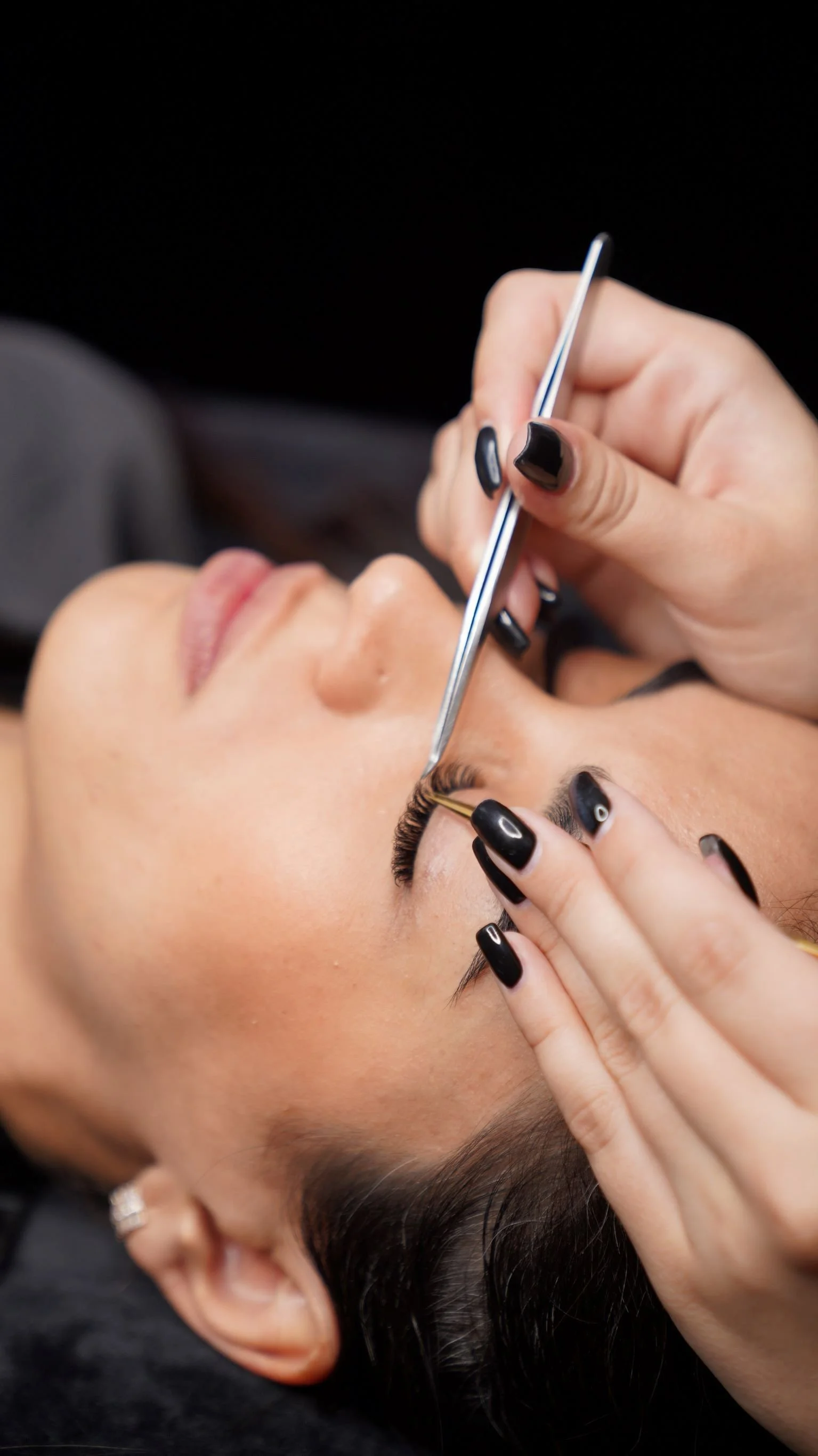 Woman receiving eyelash extension procedure with a technician applying as she reclines with her eyes closed.