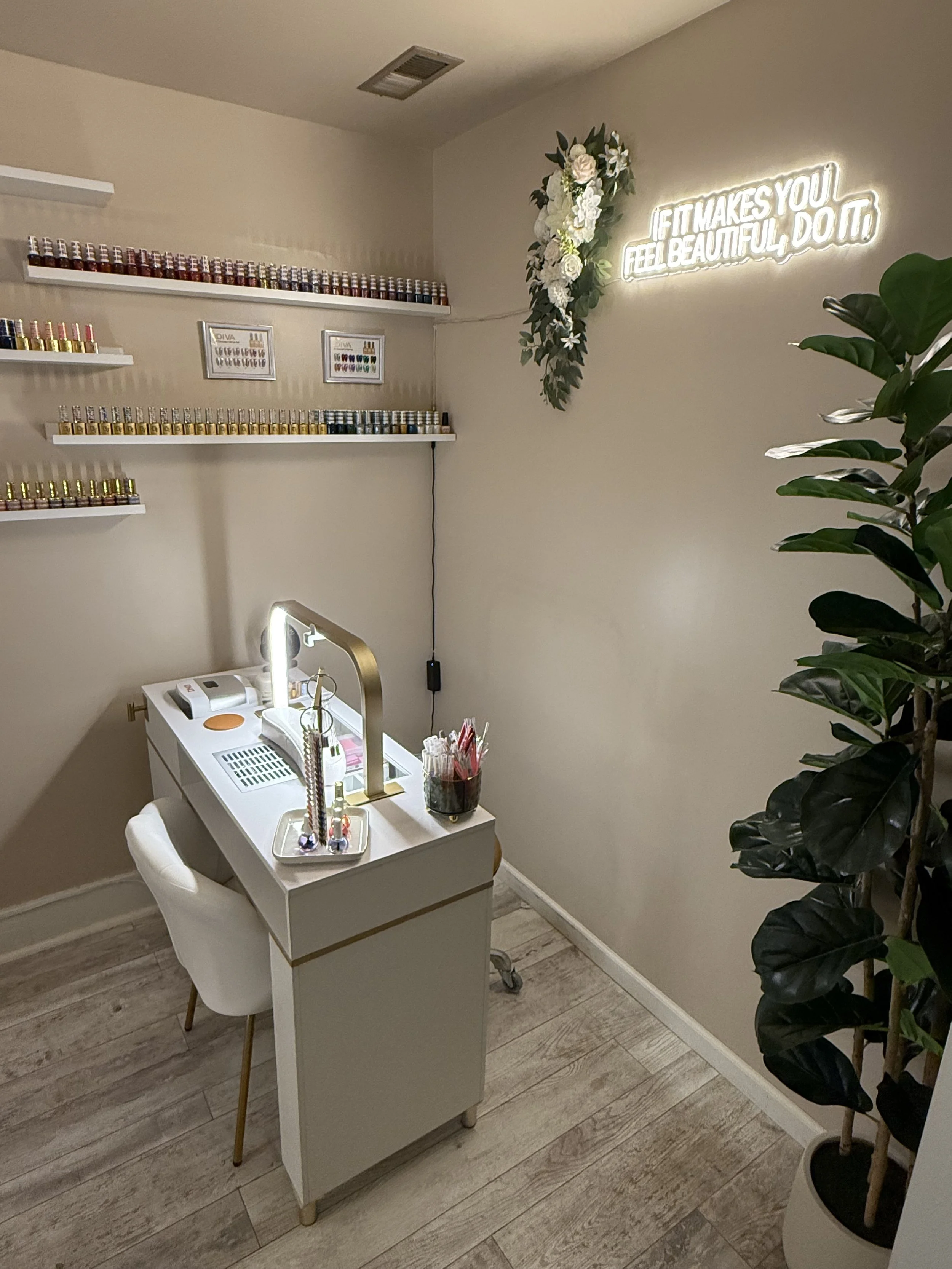 Nail salon with shelves of nail polish, a small table with a lamp, tools, and a chair, a wall with a neon sign that says 'If it makes you feel beautiful, do it', and a large potted plant.