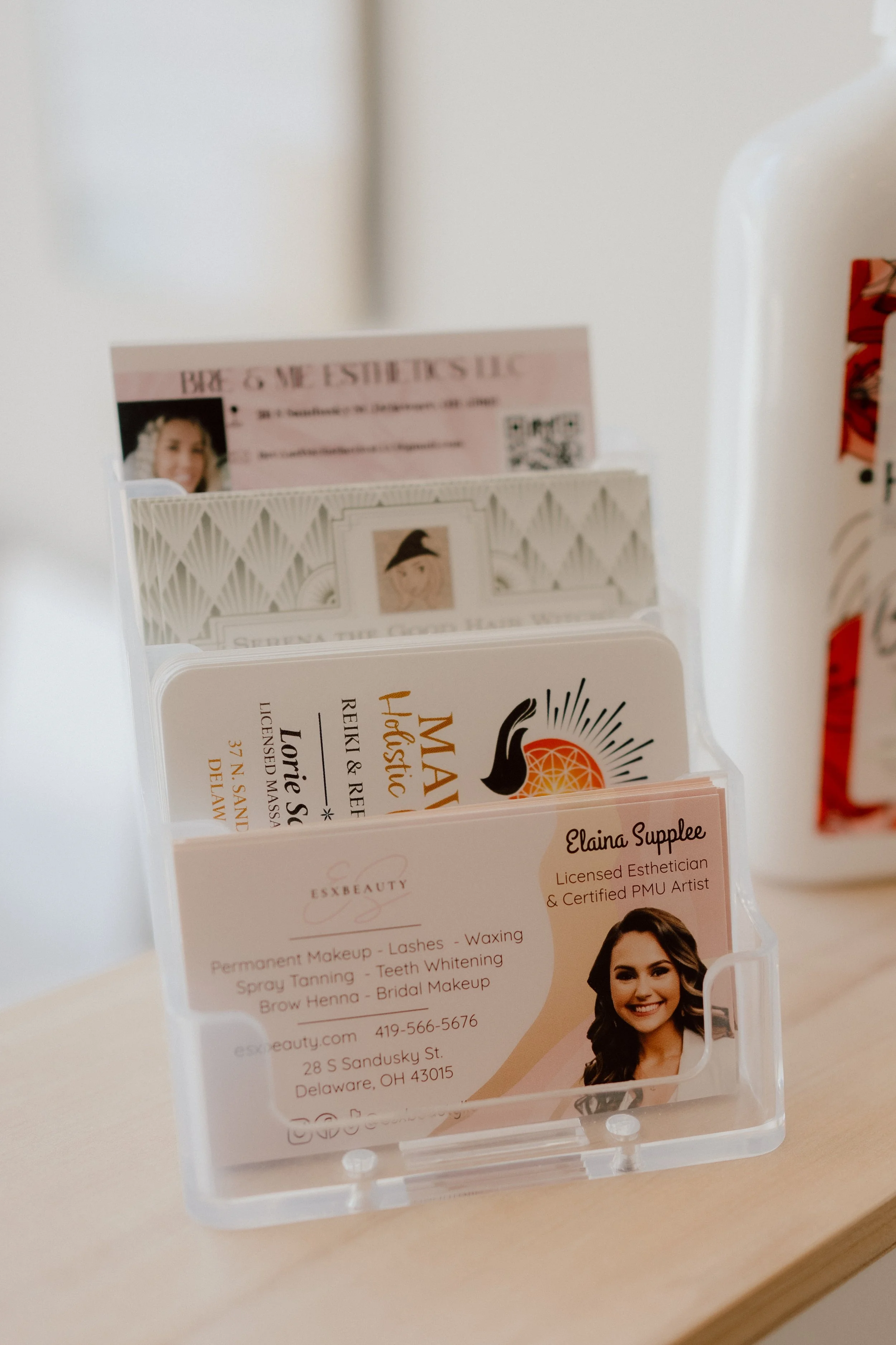 Business card holder with multiple cards, featuring a woman’s photo, business name, services, contact information, and social media icons. A jar with a red and white label is partially visible on the right.
