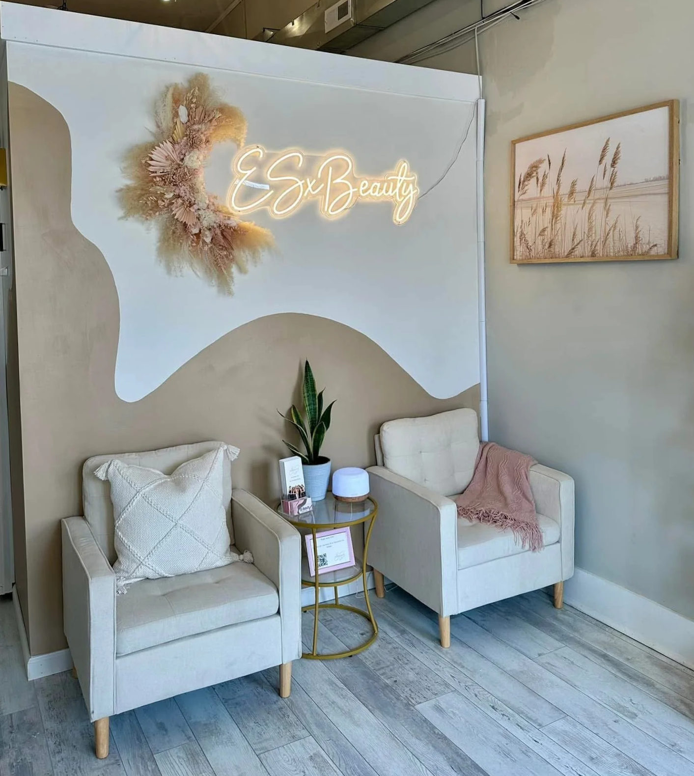 A cozy waiting area with two white armchairs, one with a decorative pillow and the other with a blush pink throw blanket. Between the chairs is a small round gold side table holding a potted plant, a name tag, and a diffuser. The wall behind has a wavy beige accent with a floral arrangement, neon sign reading 'ESxBeauty', and a framed photo of grasses by the ocean.