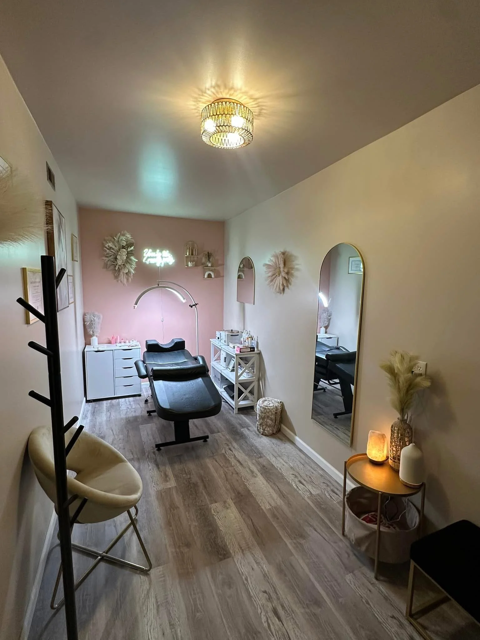A cozy spa or treatment room with a black treatment bed, a small white cabinet, mirror, decorative wall art, and soft lighting.