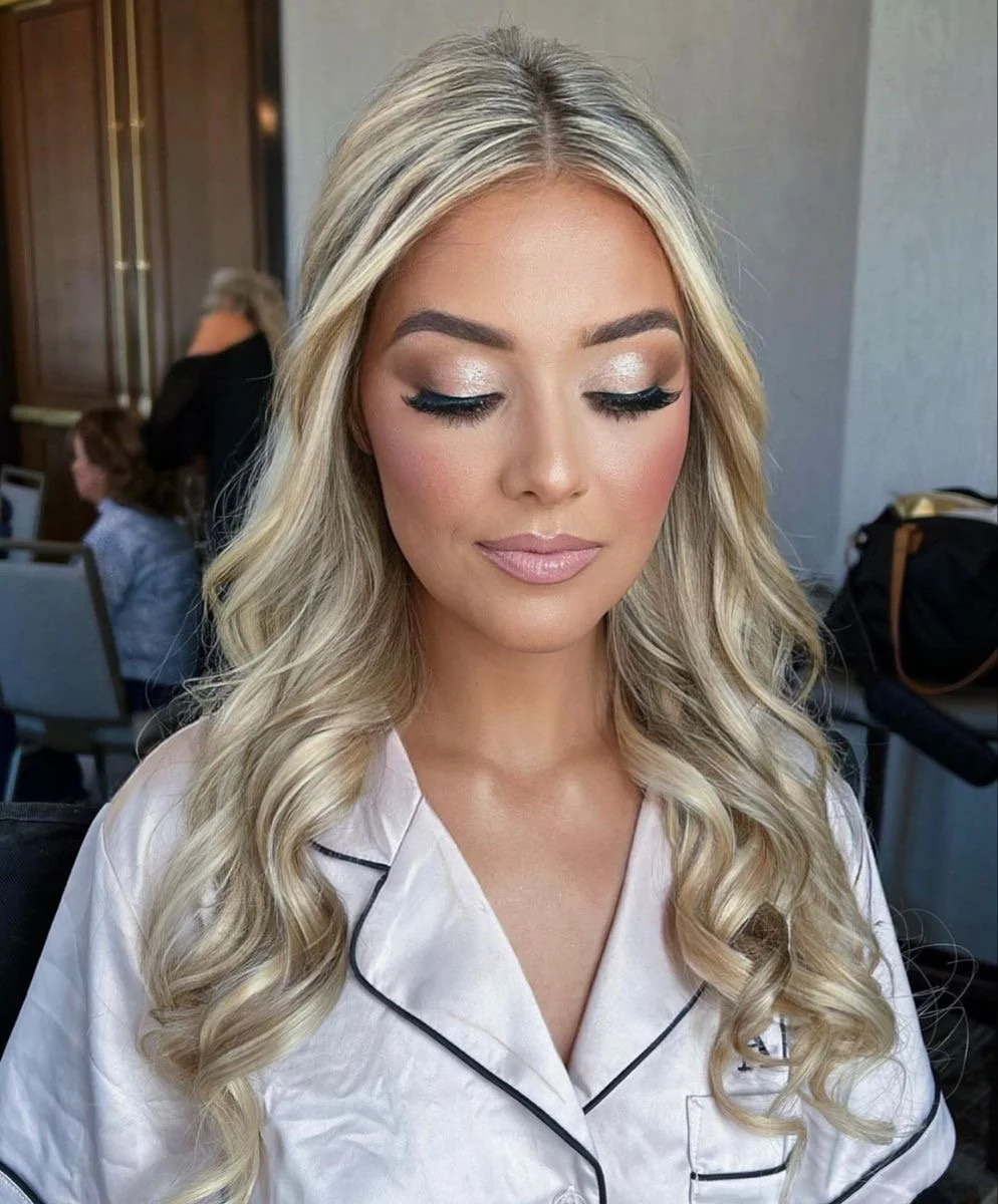 A woman in a wedding dress getting her makeup done outdoors by a makeup artist holding a makeup brush and a palette.