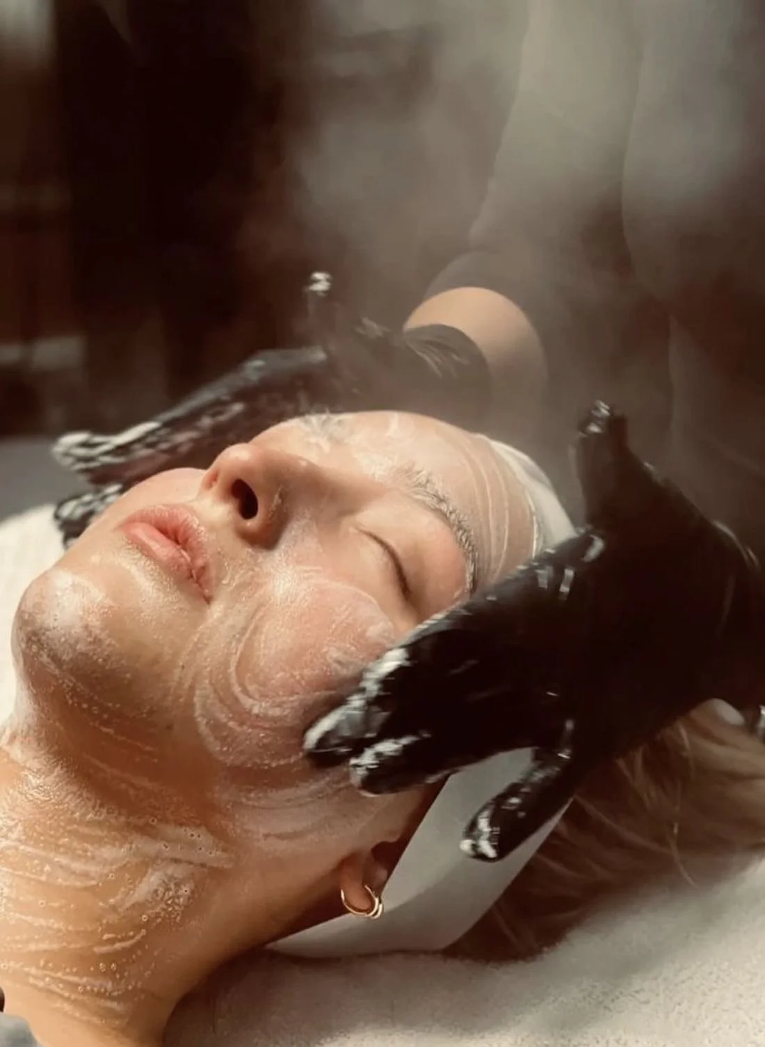 A woman lying down with a facial mask being applied by a spa technician using a brush.
