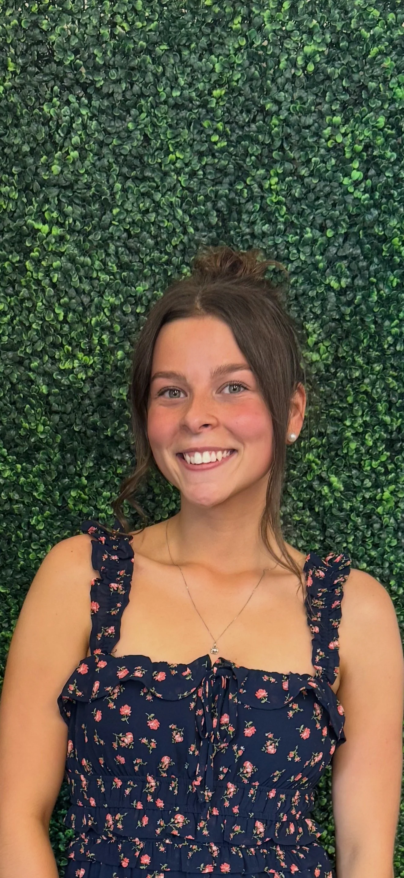 A young woman with brown hair styled in a bun, wearing a navy blue floral dress with ruffled straps, standing in front of a green leafy wall.