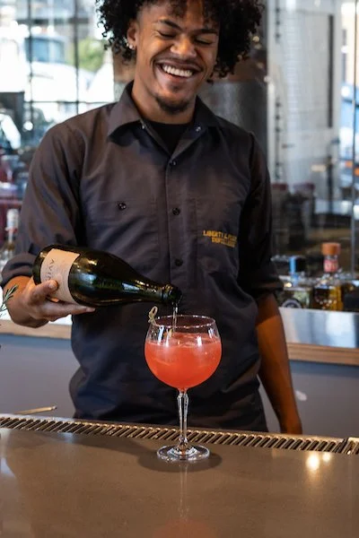 A bartender with curly hair pouring a pink cocktail into a glass at a bar.