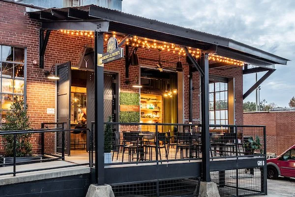 Exterior of a brick restaurant with a black metal porch, string lights, and several tables and chairs outside.