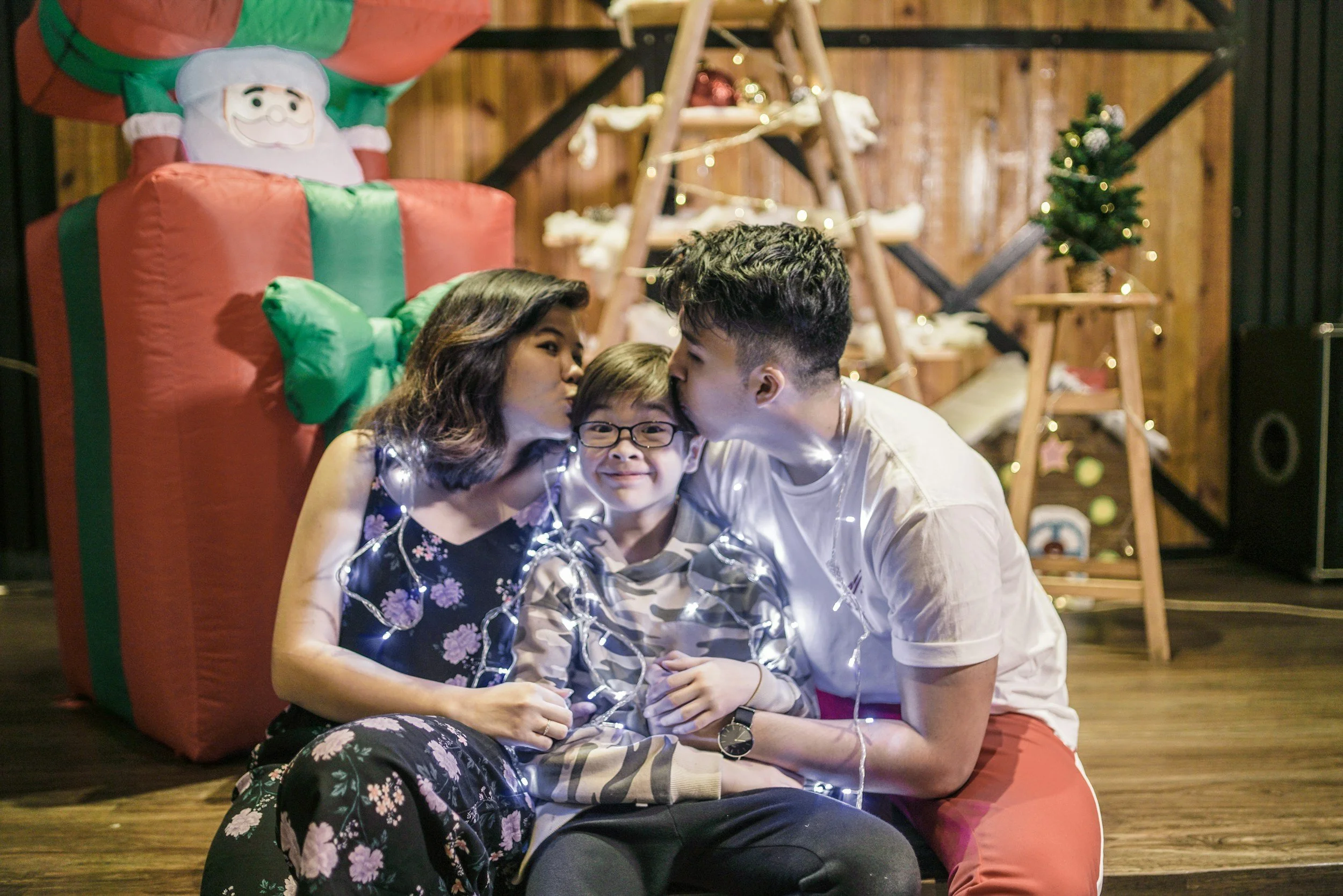A family of three sitting on the floor at a Christmas party, surrounded by holiday decorations including a large gift box with a Santa face on top, fairy lights, and a small decorated Christmas tree.