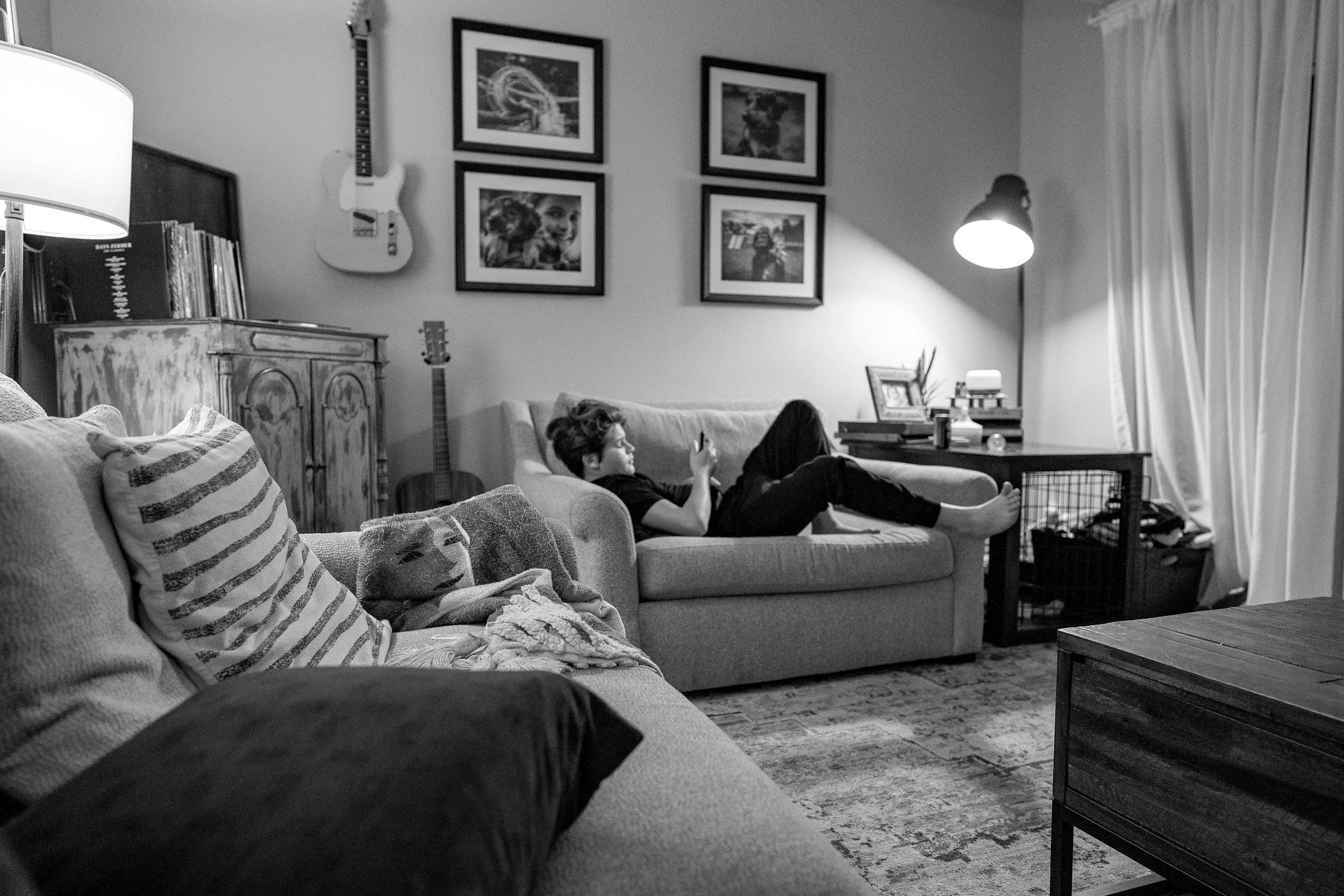 A black and white photo of a teen laying on a couch in a living room. He is looking at his phone.