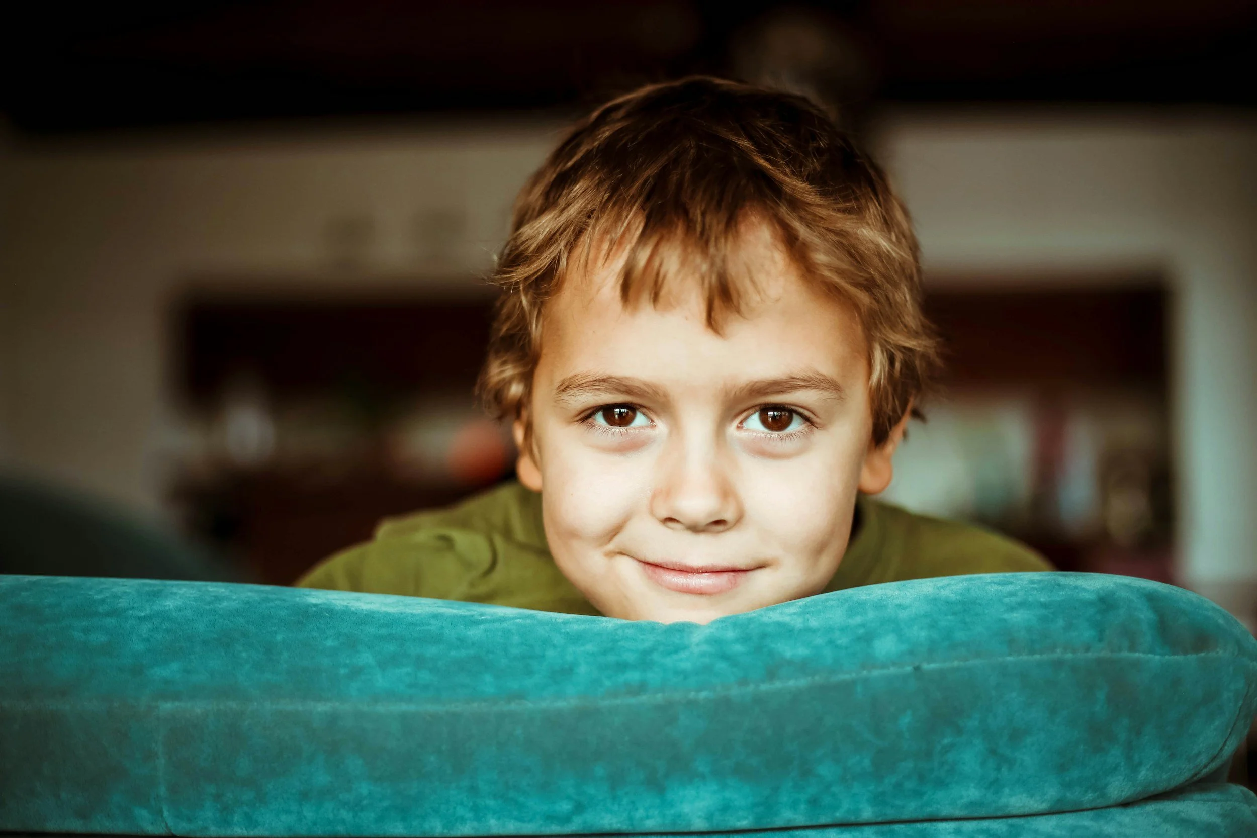 A young boy peeks his head out from over the top of the couch, he has a small smile.