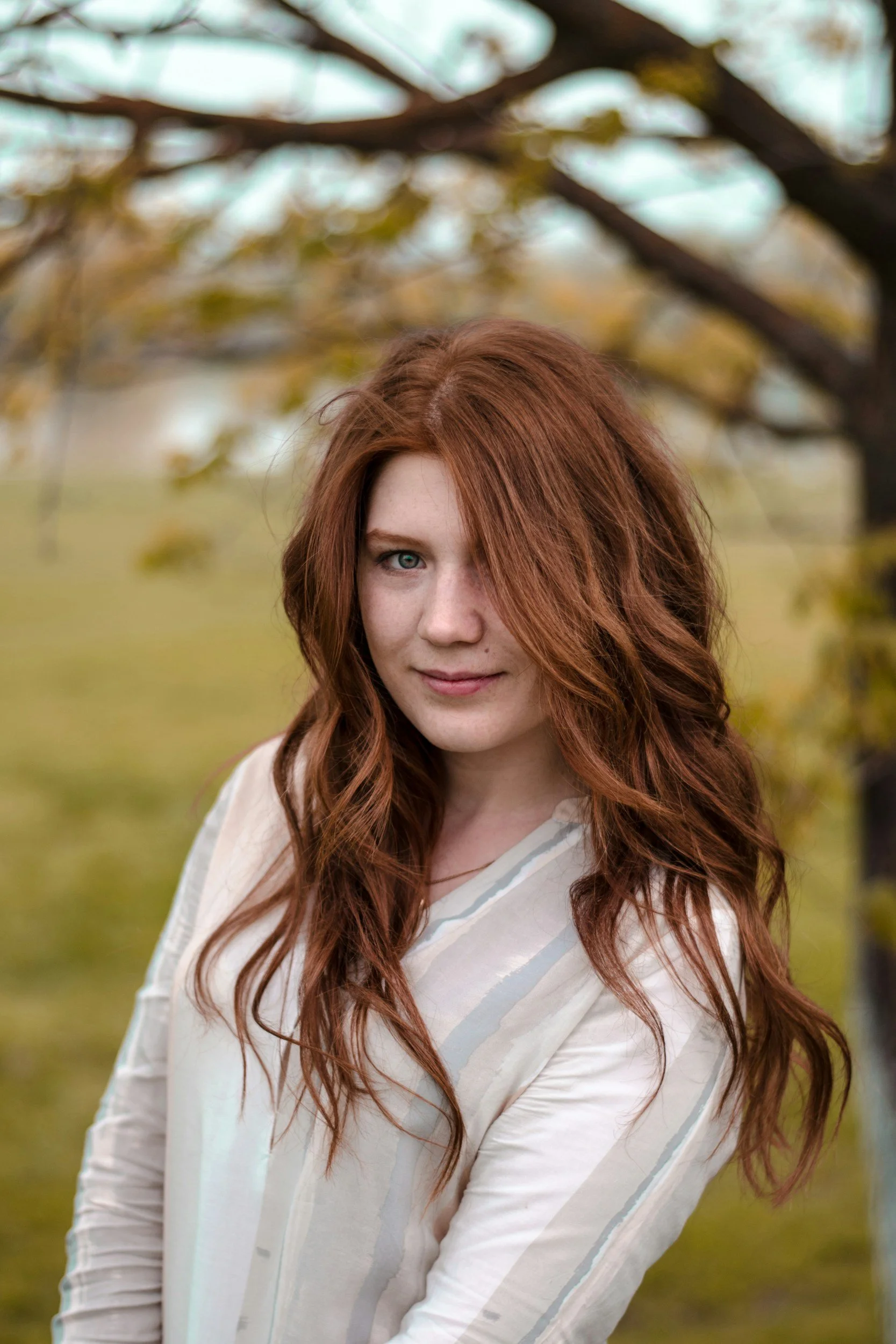 Teen Girl stands under a tree. She smiles softly, she has red hair that is shoulder length. The background contains a green field 
