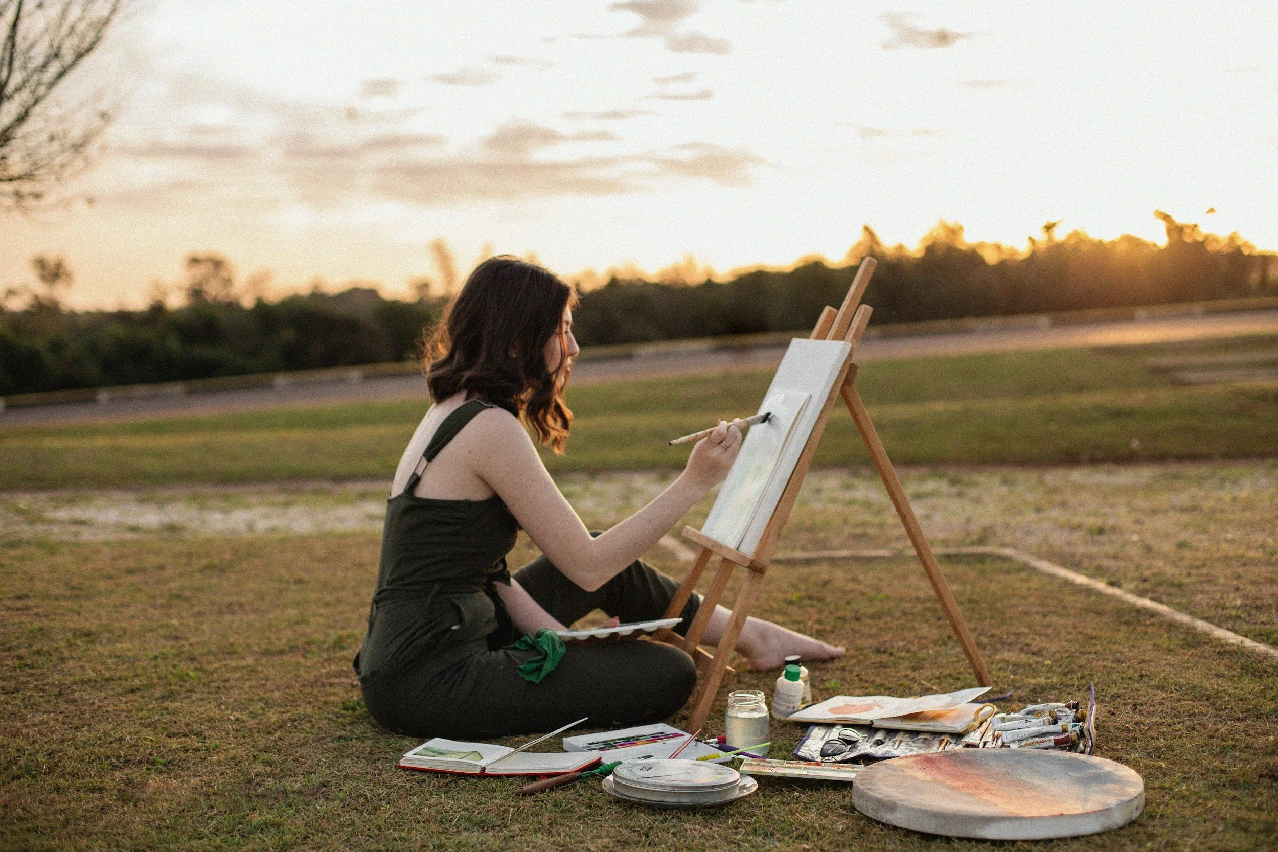 A woman paints outside. She is sitting in the grass with an easel and a variety of paint supplies. The sun is setting in the distance in the trees.