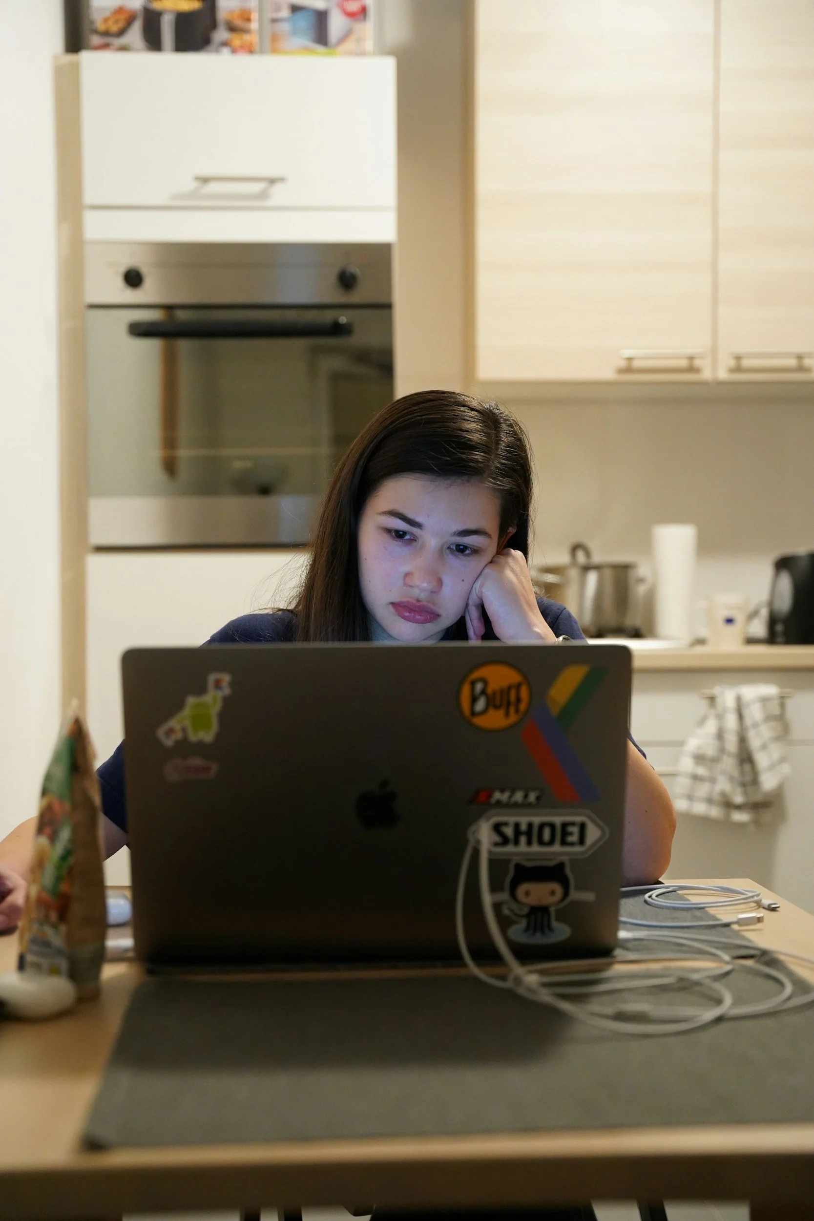 A woman sits at a lap top. Her head is resting on her hand. She looks stressed. She is in a kitchen, on the counter behind her you can see an assortment of kitchen items and a towel