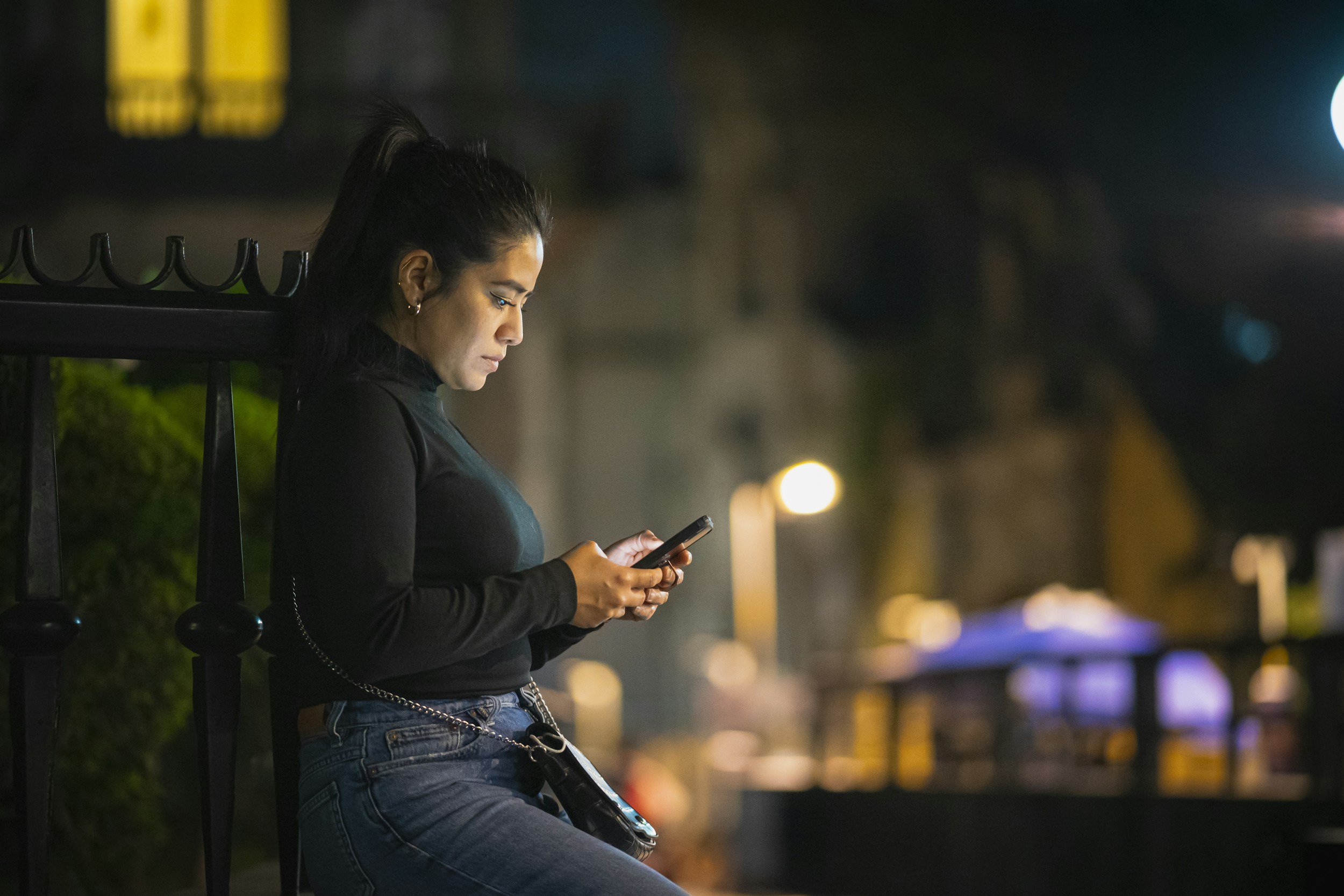 A woman is leaning against a fence, the city lights are visible in the background. She has a serious expression and she is looking at her phone.