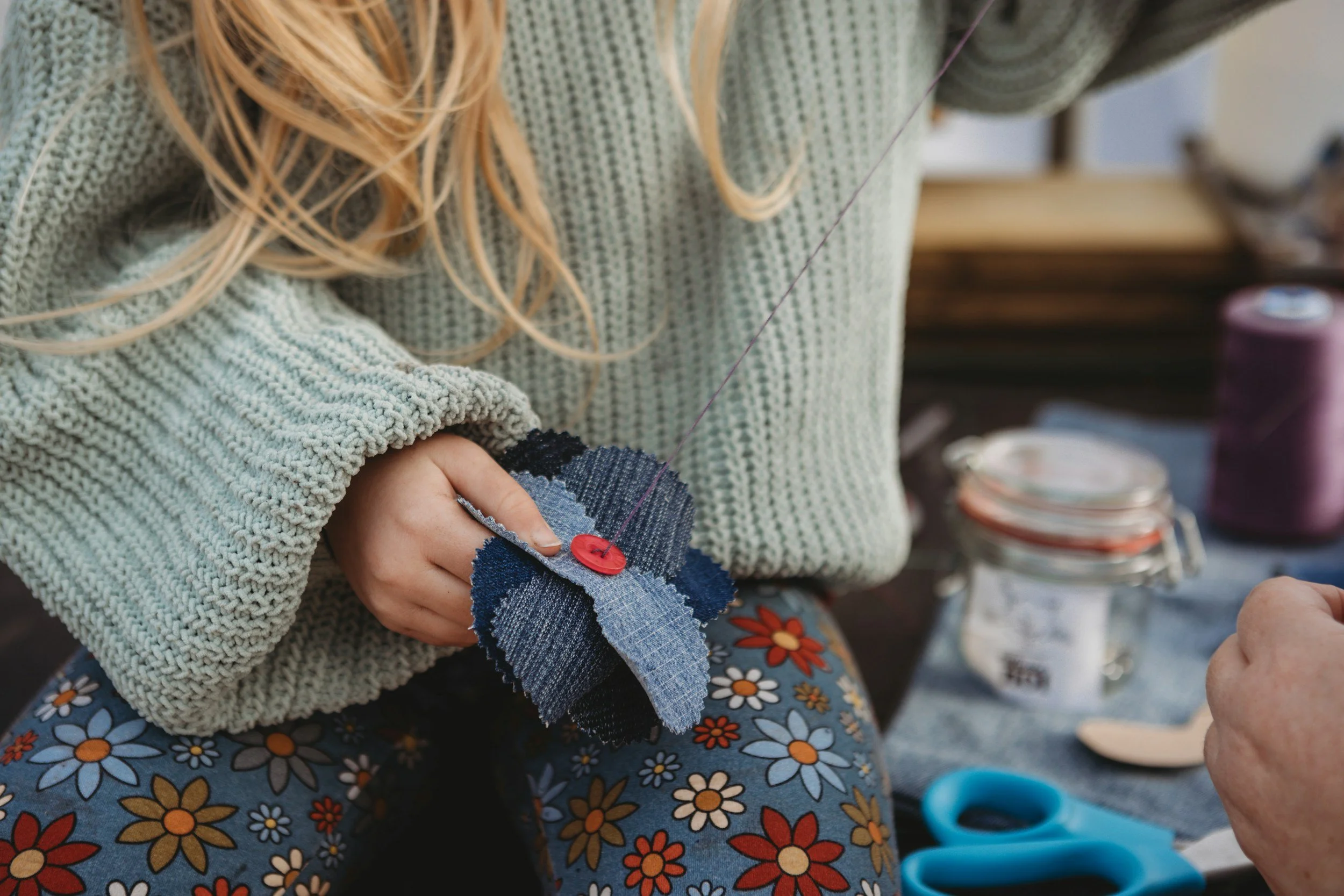 A child works on a sewing project. Her hands can be seen pulling a thread through fabric